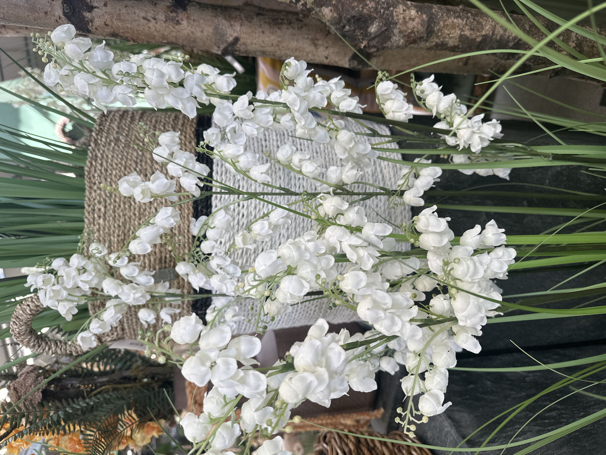 A close-up, eye-level vertical shot captures a bouquet of white artificial lily of the valley flowers arranged in a woven basket. The flowers, with their delicate bell-shaped blooms and small green buds, cascade down in clusters from slender green stems. In the background, a larger woven basket, also containing what appear to be artificial green plants, is partially visible. To the right, a textured tree trunk with vertical striations adds a natural element. The foreground is dominated by the lush green stems and leaves of the lily of the valley, with some blades of artificial grass extending upwards. The lighting is soft and even, highlighting the white flowers against the varied textures of the baskets and the tree trunk. The scene appears to be an indoor display, likely in a shop or home, possibly in Oberndorf, Austria, given the floral arrangement and natural elements.
