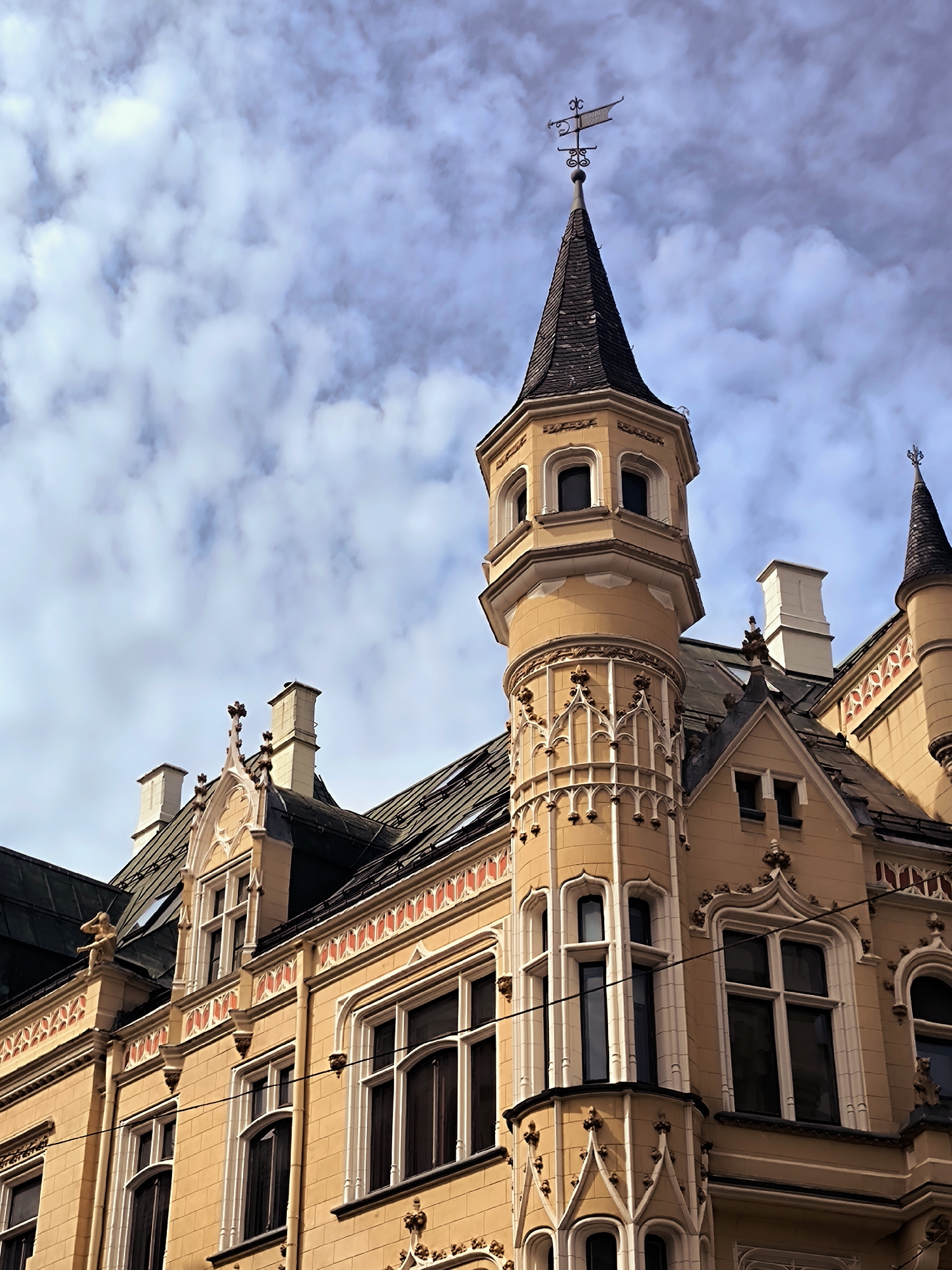 The image shows a close-up, low-angle view of the ornate facade of a historic building in Riga, Latvia. The building is predominantly a warm yellow ochre color, with detailed architectural elements. A prominent feature is a conical tower topped with a weather vane, and further down the building, another smaller conical spire is visible. The windows are framed with decorative white tracery, some with arched tops. The roofline is dark, likely slate or tile, and features several chimneys. The sky above is a mix of blue with soft, scattered white clouds. The architecture suggests a late 19th or early 20th-century Art Nouveau or historicist style, typical of Riga's historic center.