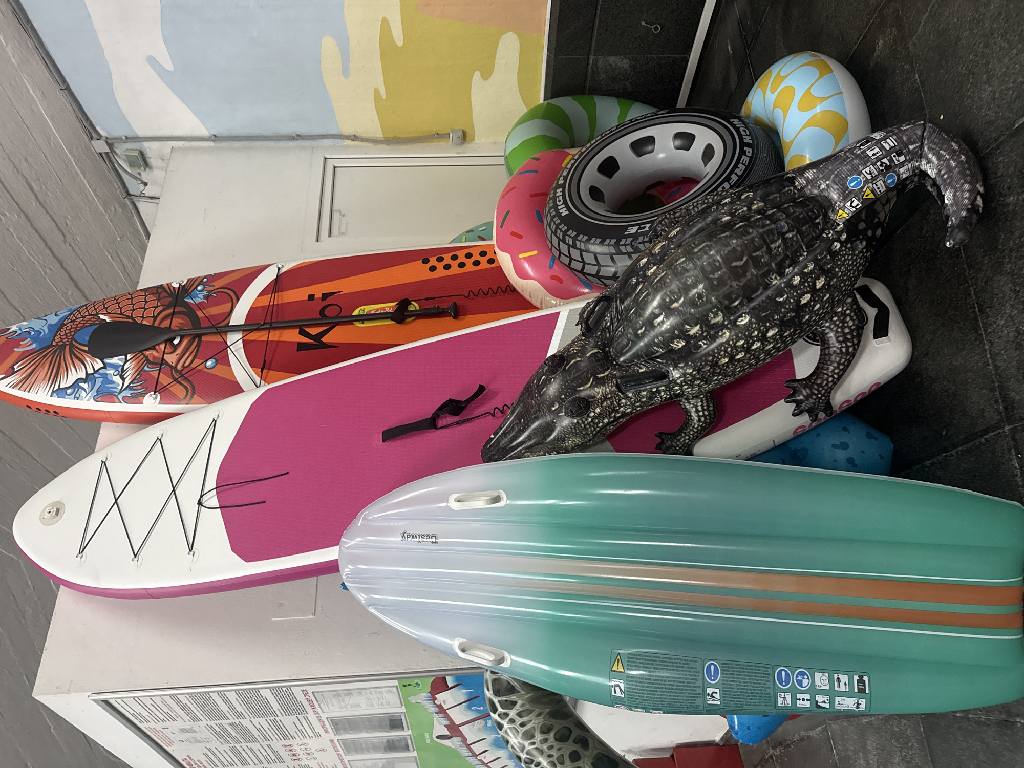 The image shows a collection of inflatable items, including two paddleboards, one pink and white and the other red with a koi fish design, leaning against a white wall. A green and white inflatable paddleboard lies horizontally in the foreground, partially obscuring other items. To the right, an inflatable alligator is positioned in front of the paddleboards. Several inflatable rings are scattered around, including a pink donut-shaped one, a green and white striped one, and one designed to look like a car wheel. A yellow and blue patterned inflatable ring is visible at the bottom right. The background includes a mural of clouds and yellow sand, and a sign with text and symbols is mounted on the white wall. The floor appears to be dark gray tile. There are no people visible in the image. The scene is indoors, likely a storage area or display for water sports equipment. The context implies a location in Yalta, Ukraine, potentially a shop or warehouse.