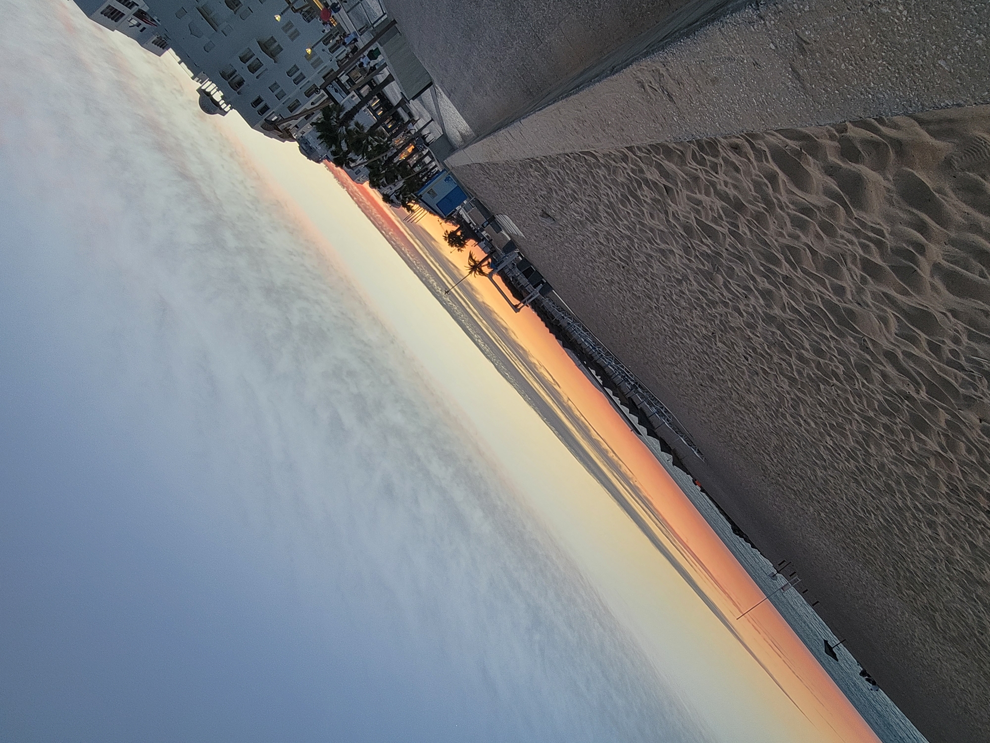 This is a tilted, low-angle photograph of a beach in Quarteira, Portugal, during what appears to be sunset or sunrise. The sky transitions from a clear blue at the top left to soft hues of orange and pink closer to the horizon line, with wispy clouds present. The sea is visible in the distance, bordering the beach. The sandy beach occupies the majority of the foreground and midground, showing textured sand patterns. Along the right side of the image, there is a concrete embankment and walkway, with buildings and palm trees visible beyond it. Several beach chairs and umbrellas are set up on the sand. A few people are faintly visible on the beach and on the walkway.