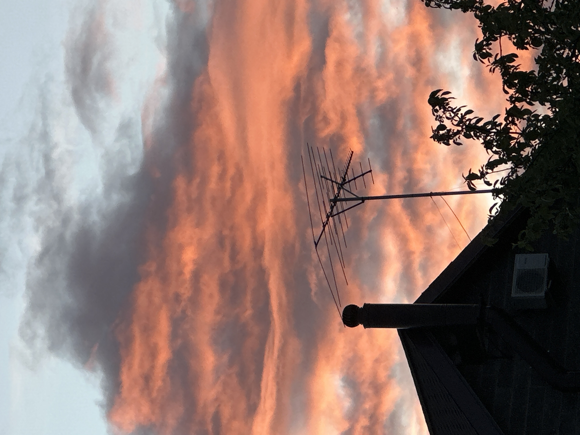 The image shows a view of a house with a chimney and antenna silhouetted against a vibrant, cloudy sky at sunset or sunrise. The sky is filled with hues of orange, pink, and gray, indicating warm light reflecting off the cloud formations. A tree with dark foliage is visible on the right side of the frame, partially obscuring the house. The house has a gabled roof and an air conditioning unit on the wall. The photograph appears to be taken from a low angle, emphasizing the height of the house and the dramatic sky. There is no information provided or apparent that reveals that the location is the city of Nizhny Novgorod, Russia.