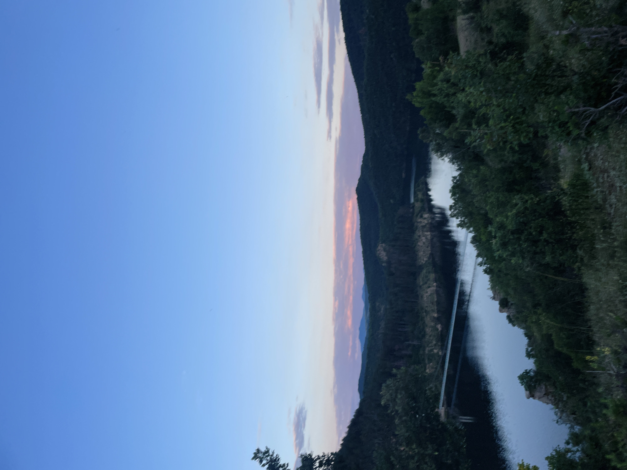 The image depicts a serene landscape at dusk, likely near the city of Suhuvo, Bulgaria. A body of water, possibly a lake or reservoir, is visible, reflecting the soft hues of the twilight sky. A long, slender bridge spans across the water, supported by visible piers. The surrounding terrain is characterized by rolling hills covered in dense forestation, with trees creating a dark silhouette against the fading light. In the distance, a range of mountains can be seen, their peaks softened by the atmospheric haze. The sky transitions from a deep blue at the top to lighter shades of pink and lavender near the horizon, where wispy clouds are present. The foreground shows a grassy, uneven slope dotted with vegetation, suggesting a vantage point overlooking the scene. There are no people or animals visible in the image.