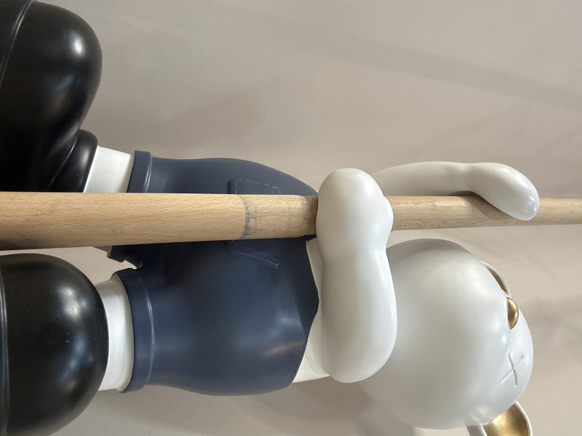 The image shows a vinyl art toy figure of a character with a round white head, gold ears and facial features, and x-shaped eyes. The body is mostly navy blue, with white sections connecting to the large black spherical feet. The figure is gripping a light brown wooden rod with both hands, appearing to hang from it. The background is a plain off-white wall. The location and city are not identifiable from this image.