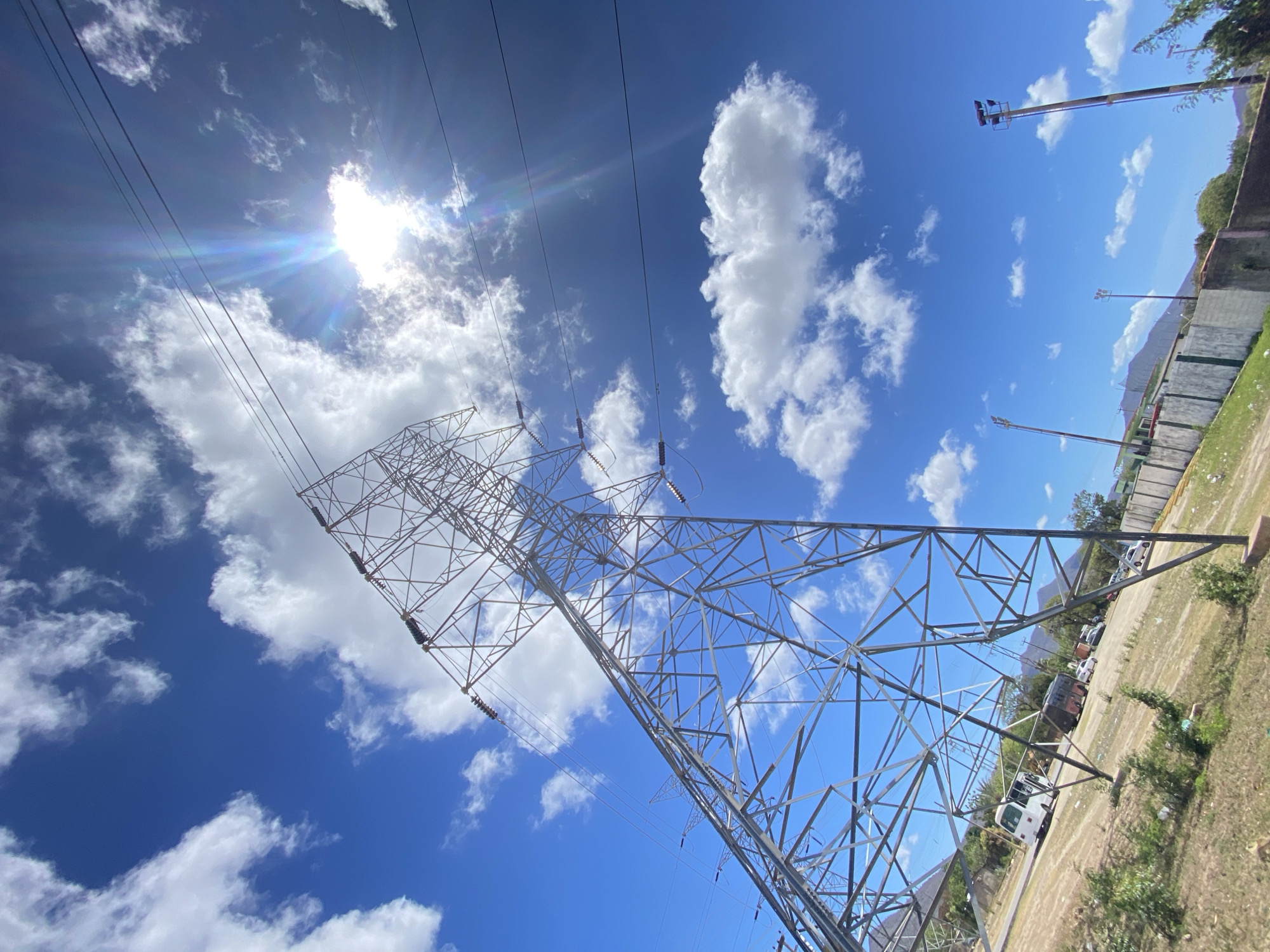The image shows a high-voltage power transmission tower in Juan Griego, Venezuela. The tower is made of a lattice of steel beams and is situated against a backdrop of a blue sky with scattered white clouds and the sun peeking through. High-tension power lines run from the top of the tower. Adjacent to the tower is a parking area with parked vehicles. A perimeter wall surrounds the property.
