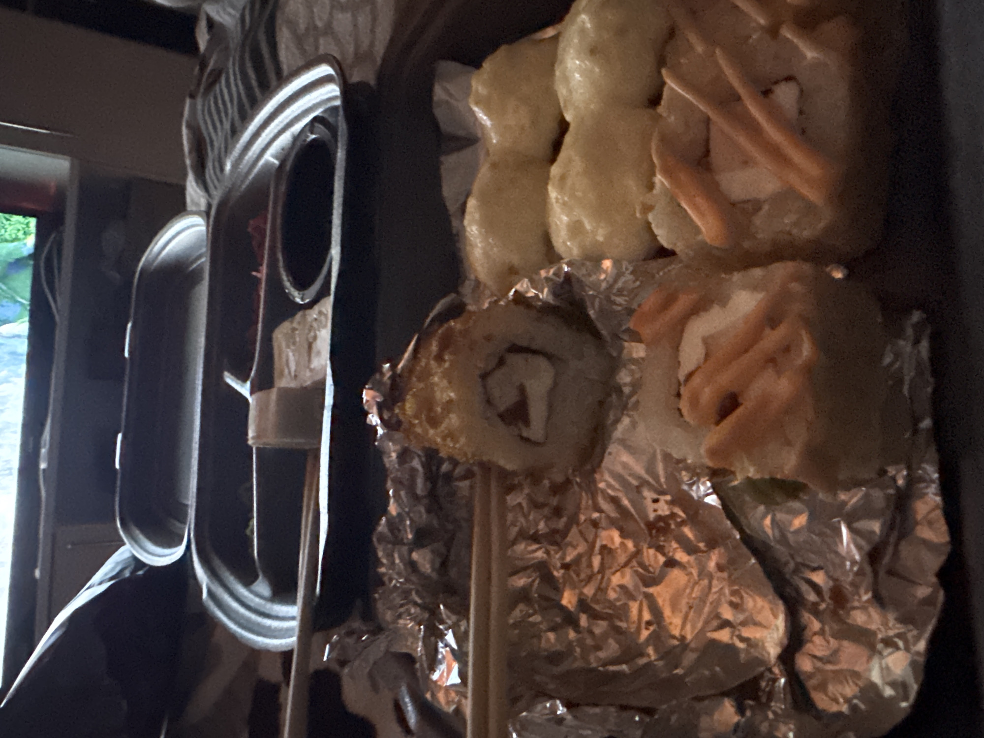 The image shows a take-out container of sushi resting on crumpled aluminum foil. A single sushi roll is impaled on a chopstick. The background shows a partial view of an interior, possibly a home or restaurant. The presence of the sushi does not inherently locate the scene in Izhevsk, Russia, or any specific city.