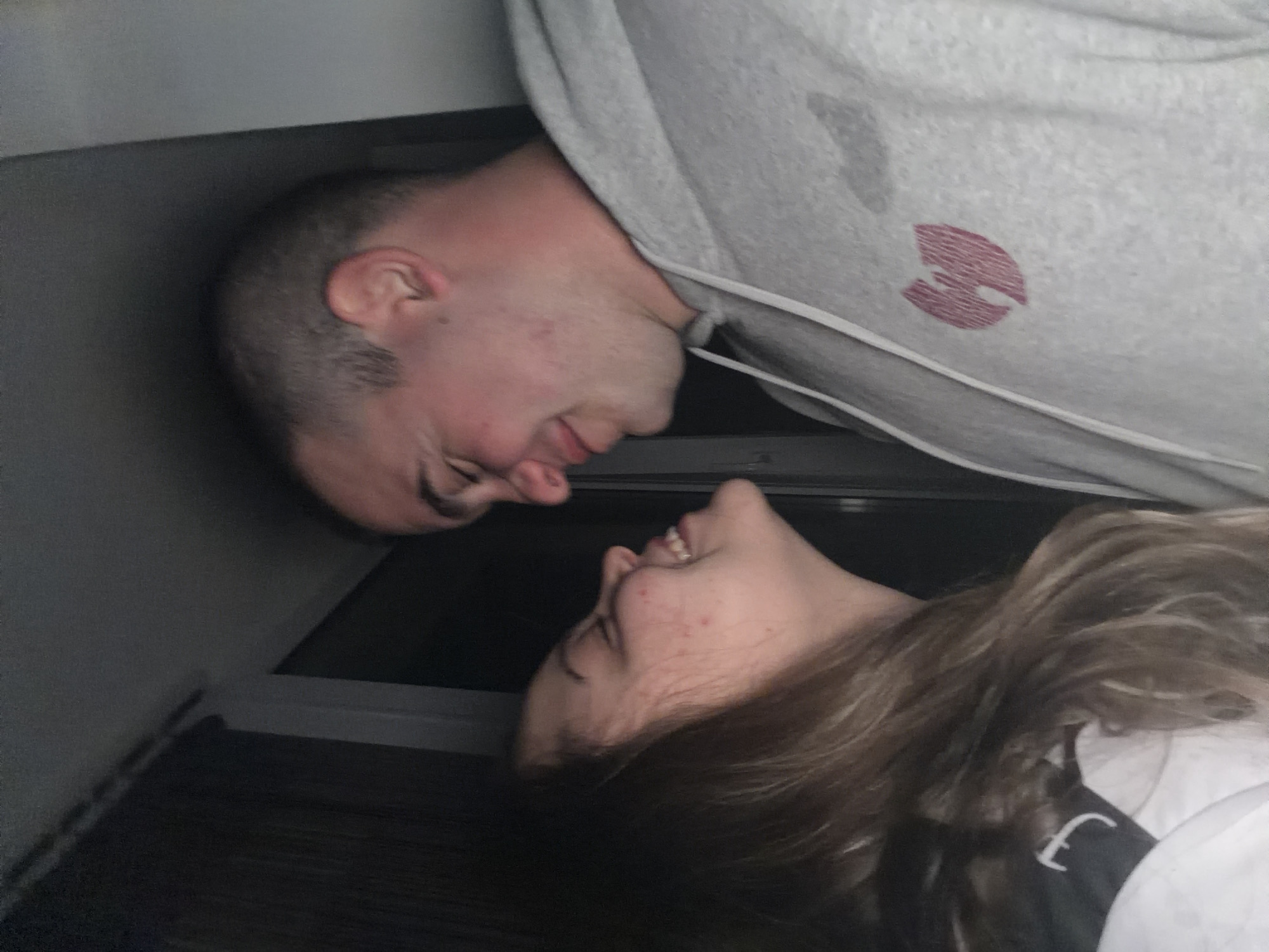 The image, rotated 90 degrees clockwise from its original orientation, captures an intimate moment between two young individuals in what appears to be a dimly lit indoor setting. A male and a female are seen face-to-face, their heads tilted towards each other, suggesting a tender connection, possibly on the verge of a kiss or sharing a close moment. Both subjects are smiling, conveying emotions of happiness and affection.
The male, positioned on the left side of the frame (when viewed upright), has short, dark hair and a light complexion with some facial redness. He is wearing a light grey hooded sweatshirt. A prominent dark pink or reddish lip print is clearly visible on the upper arm or shoulder area of his sweatshirt, indicating a kiss.
The female, positioned on the right, has long, wavy brown hair that frames her face and falls over her shoulder. She is seen smiling widely, revealing her teeth, and has some natural blemishes on her cheek and forehead. She is wearing a dark top with a white, stylized symbol, possibly an 'F' or part of a logo, visible on her left shoulder area.
The background is dark and largely indistinct, featuring horizontal lines that could belong to an interior structure like a seat or wall panel, suggesting a confined space such as a vehicle or a compact room. The scene evokes a sense of personal connection. The location context is Varna, Bulgaria.