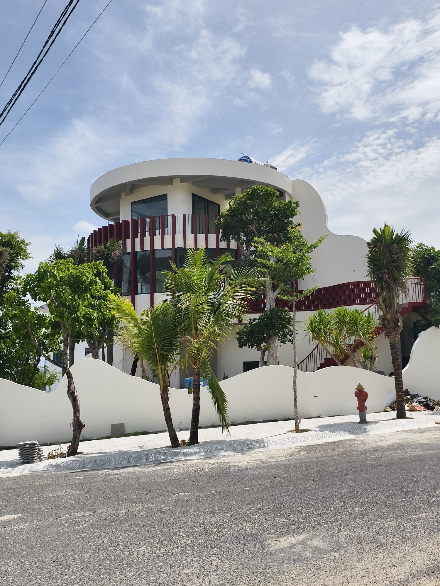 The image shows a modern, two-story building with a cylindrical upper level, located in Bac Nha Trang Ward, Vietnam. The building is white with red vertical accents on the second story and red curved stairs on the side. A wavy white wall surrounds the property, with a few trees and palm trees visible within the wall. A red fire hydrant is located on the sidewalk near the road. The sky is partly cloudy, with power lines visible in the upper left corner. The road is asphalt and appears to be lightly traveled. A stack of tiles or concrete pavers are on the sidewalk to the left. Debris is on the ground behind the fire hydrant.