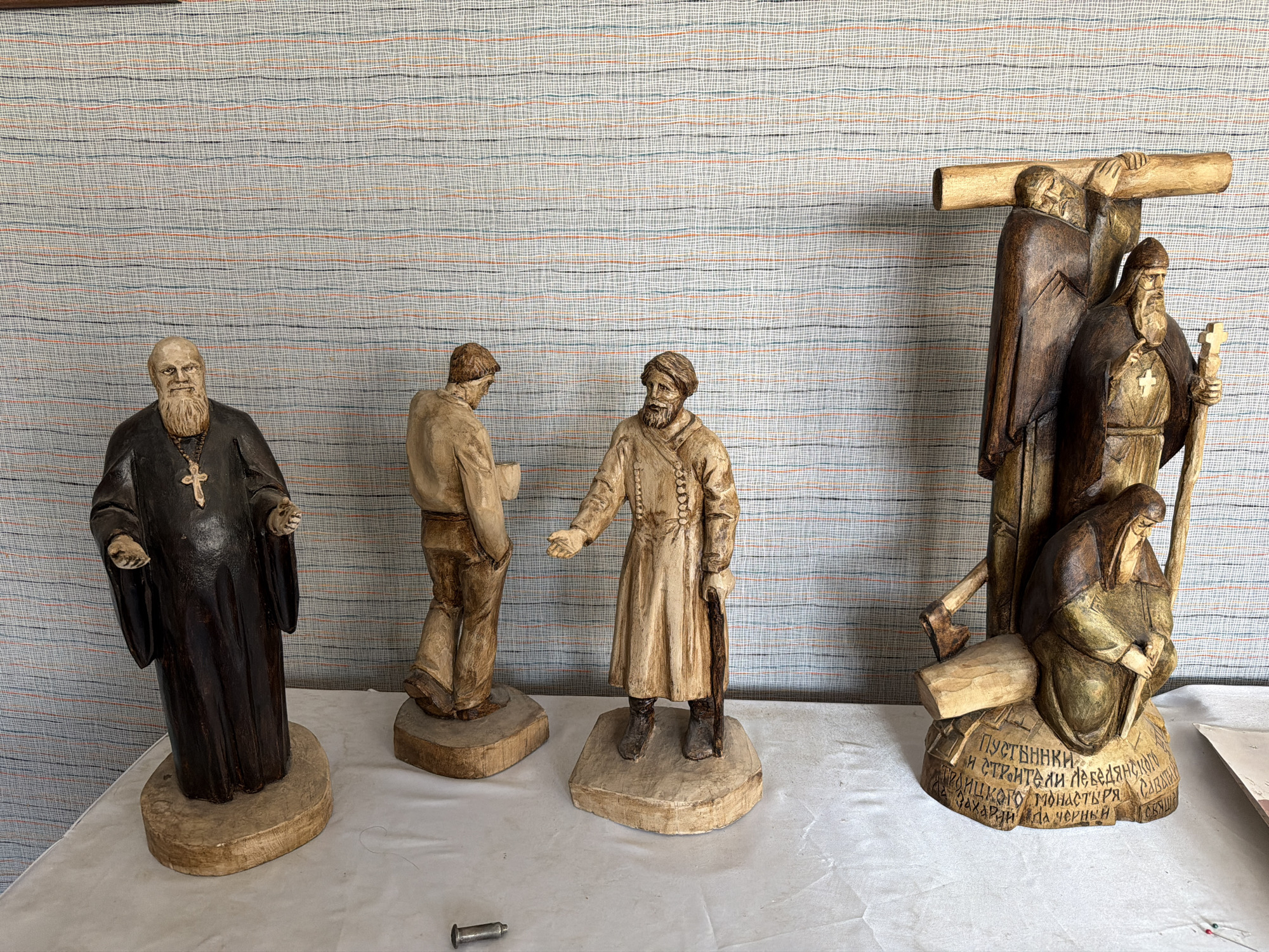 The image shows a collection of wooden sculptures displayed on a white tablecloth against a striped wallpaper background. The sculptures depict religious figures and scenes, with intricate carvings and a rustic aesthetic. On the left, a robed figure with a beard and a cross pendant stands prominently. Next to him, two other figures are carved in more plain attire. To the right, a larger sculpture features a group of figures associated with a cross, possibly representing a biblical scene. The base of this sculpture is inscribed with Cyrillic text. The background wallpaper has a repeating pattern of thin horizontal lines in shades of blue, orange, and black. A metal object, possibly a screw or fastener, is visible on the tablecloth in the foreground. The overall impression is of a religious art display, likely from a local craftsman or religious institution. The text on the base of the sculpture on the right refers to "Пустынники и Строители Лебедянского Захария monasteries," which translates to "Hermits and Builders of the Lebedyan Zakharia Monastery," indicating the location of origin in Lebedyan, Russia.
