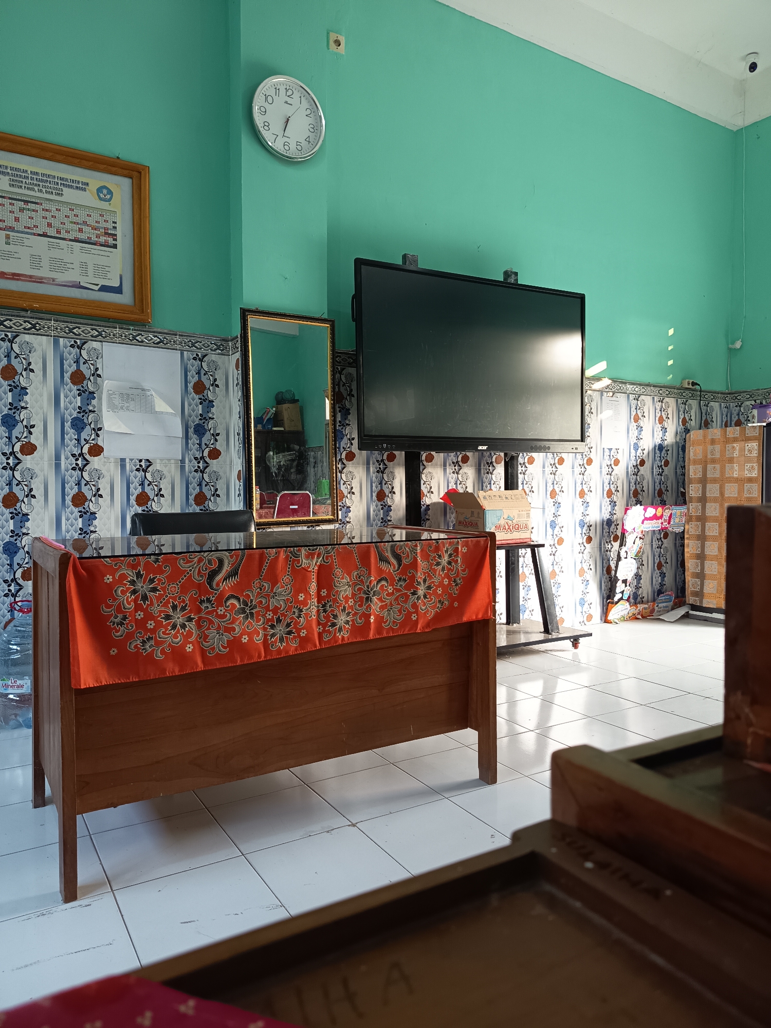 The image depicts an interior scene, likely a classroom or administrative office, in Widoro, Indonesia. The walls are painted a vibrant turquoise, and the floor is tiled with white squares. To the left, a framed document with Indonesian text is mounted on the wall. Below it, a patterned tile backsplash runs horizontally.
Dominating the center of the frame is a wooden desk with a glass top, draped with an orange cloth featuring a black and white floral pattern. On the desk, a large mirror with a gold frame is propped up, reflecting a red chair. A large flat-screen television is mounted on a stand behind the desk. To the right, a display rack holds various items, and further to the right, a patterned wall adorns the space.
The lighting suggests it is daytime, with sunlight casting shadows across the floor and illuminating parts of the room. There are no people visible in the image, and the overall impression is one of an organized, functional space.