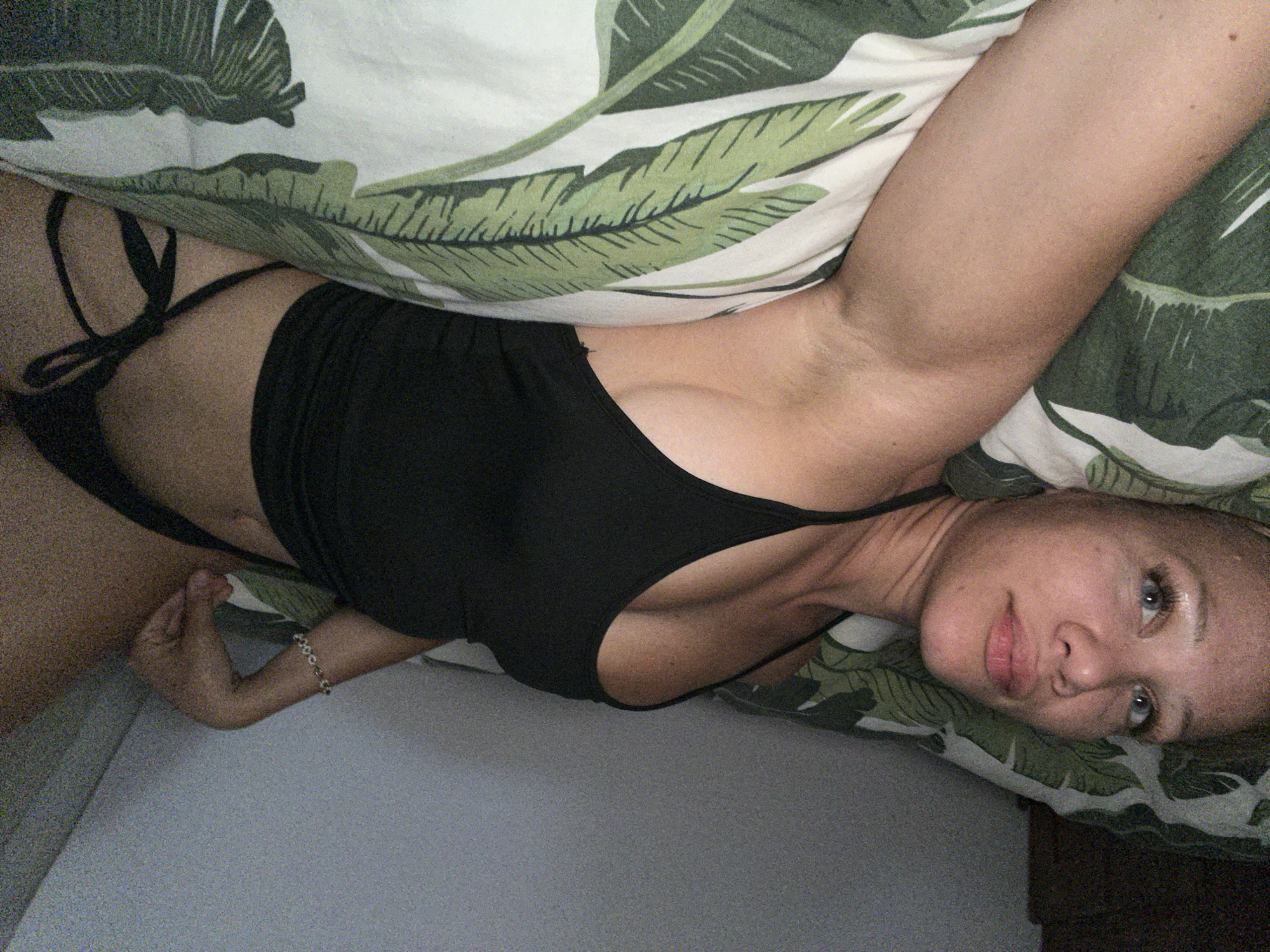 A woman with blonde hair and blue eyes lies on a bed with green and white leaf-patterned sheets. She is wearing a black crop top and black bikini bottoms. The woman is lying on her back with her head turned towards the camera. She has a bracelet on her left wrist. The scene is indoors, in what appears to be a bedroom. There is no indication of the specific location within Mühldorf a. Inn, Germany.