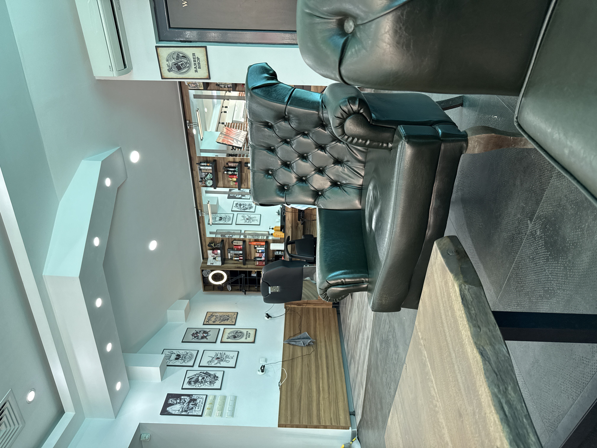 The image depicts the interior of a barber shop in Shumen, Bulgaria. The foreground features a rough-hewn wooden table and a dark green tufted leather armchair, with another similar armchair partially visible to the right. The background reveals a wall adorned with framed black and white artwork, possibly related to barbering or popular culture, and a wooden paneled section. Further back, a barber chair is visible, alongside shelves stocked with various barbering products and tools. A circular illuminated sign hangs above the shelves. An air conditioning unit is mounted high on the wall. The overall ambiance suggests a modern, stylish barbershop.