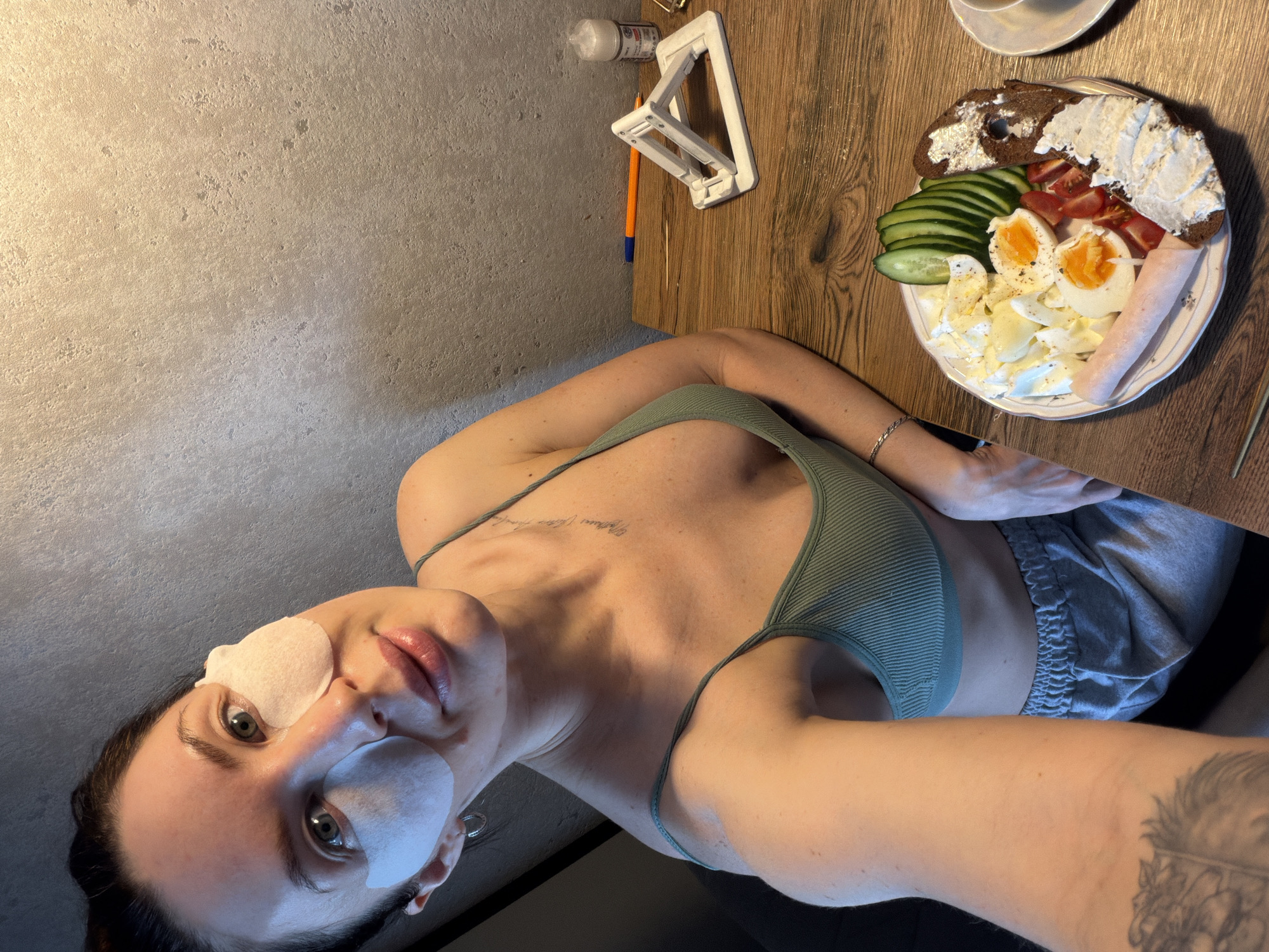 A woman is taking a selfie. She has eye patches on. She is wearing an olive green tank top and gray shorts. In the background, there is a plate of food with bread, cream cheese, cucumber, tomato, eggs, and ham. There is also a phone stand, pen, and bottle on the table. The location is likely indoors in Perm, Russia.