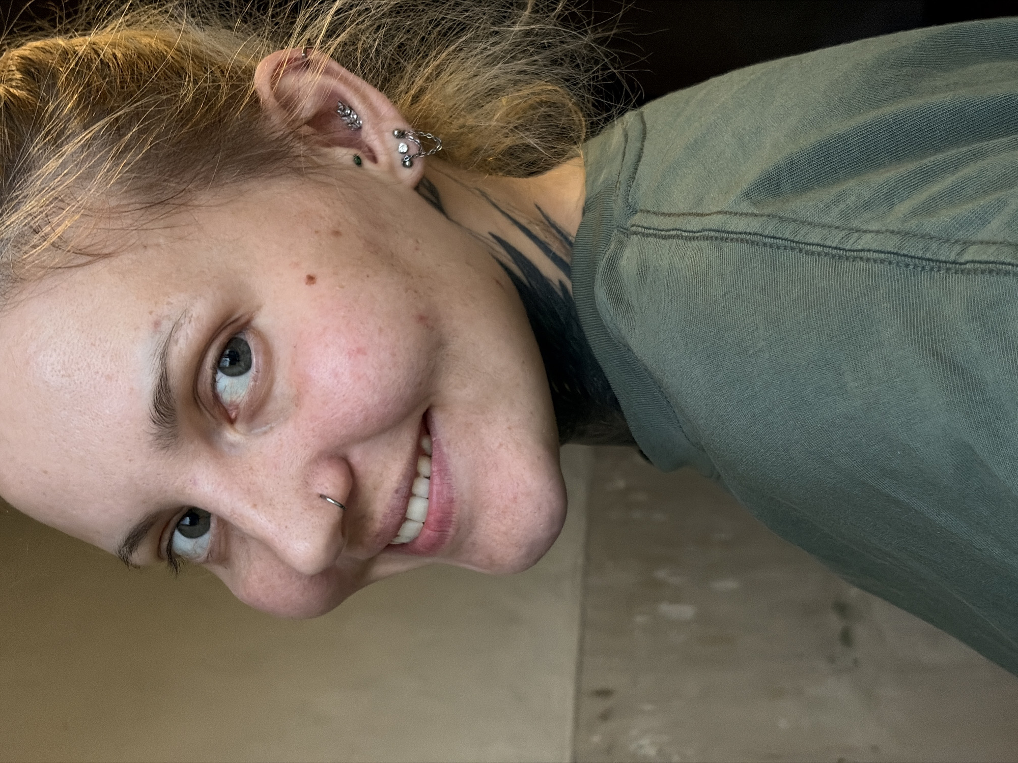 A close-up, angled headshot shows a person with fair skin, light blonde hair, and gray eyes. They have a nose ring and multiple piercings in their left ear, including a conch piercing, a helix piercing, and several lobe piercings. A dark tattoo is visible on their neck. They are wearing a green shirt. The background is slightly blurred and includes a light beige wall. The image location is not provided in the image.