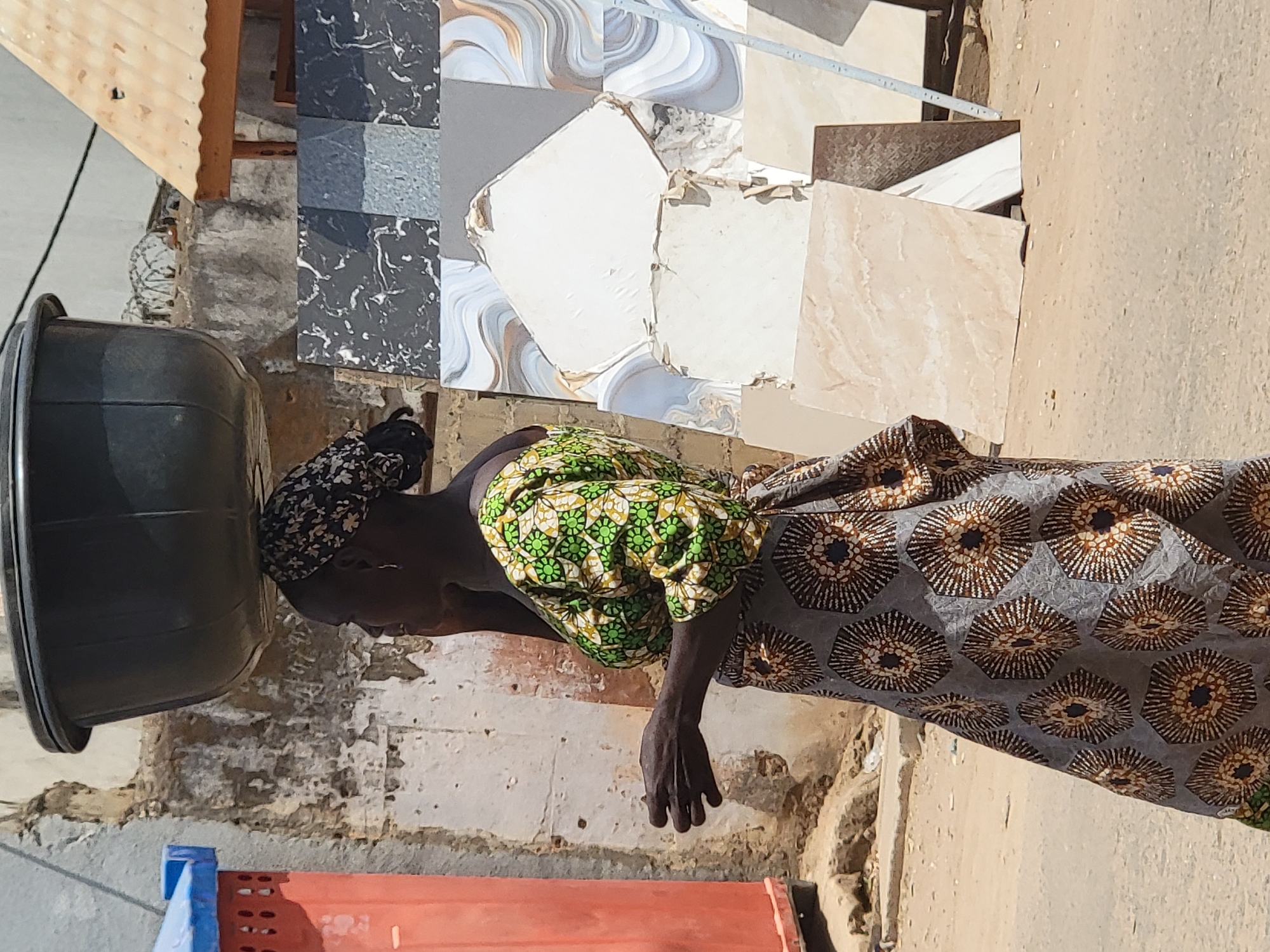 In Maiburiji, Nigeria, an individual wearing a patterned dress and head covering is positioned next to a large, dark-colored bucket. Adjacent to the person are various tiles stacked against a wall. A red and blue plastic object is visible on the ground near the bucket. The scene appears to be outdoors.