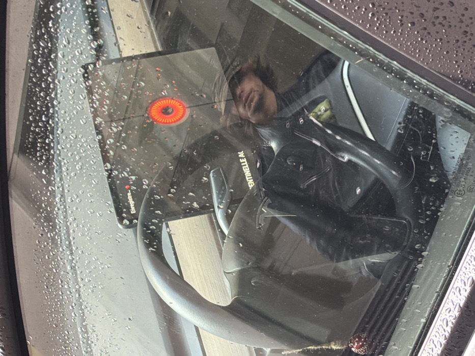 Through a rain-speckled car window, a reflection shows a person with shoulder-length hair and a black shirt. Reflected next to them is a black kitchen appliance with an orange, lit ring on its surface. The car's steering wheel is visible in the foreground, partially obscuring the reflected scene. Part of the dashboard is visible on the right side of the car window.