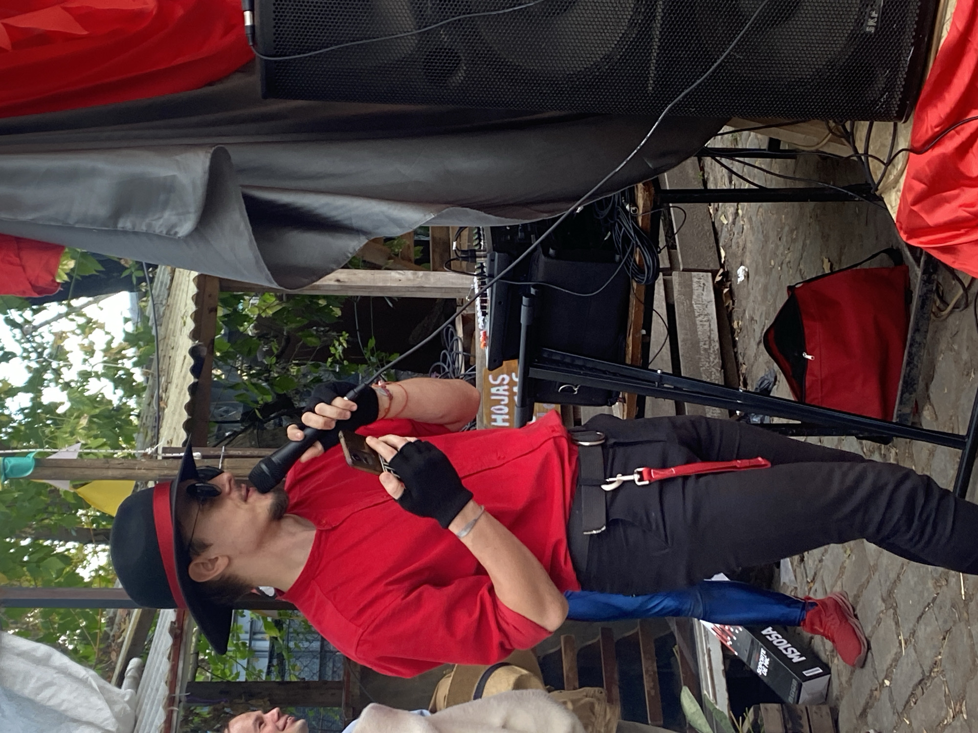 A man in a red shirt, black pants, and a black hat with a red band is holding a microphone and a smartphone. He's outdoors, seemingly on a makeshift stage or platform. There is sound equipment and a black metal structure surrounding him. There is foliage behind the structure. A glimpse of a blue item and red shoes are visible. At the bottom of the image, parts of other people are visible. The setting is likely a public space, potentially in Buenos Aires, Argentina.