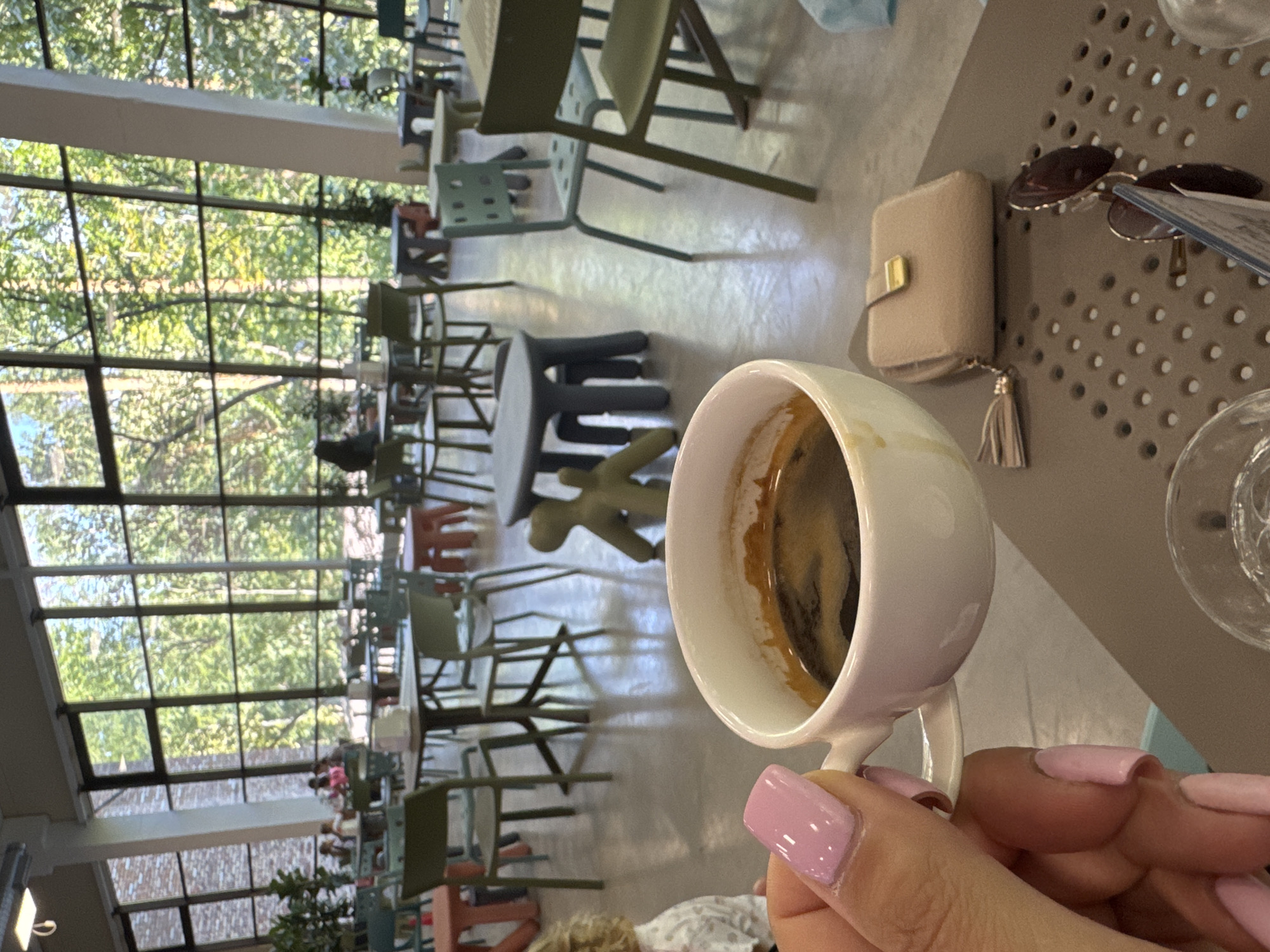 A close-up shot shows a hand with light pink manicured nails holding a white ceramic cup filled with dark coffee, with some foam visible on top. The hand and cup are positioned in the foreground, slightly to the left. Behind the cup, on a table, rests a small, light beige wallet with a gold clasp and a beige tassel. Next to the wallet, a pair of sunglasses with dark lenses and gold frames is partially visible. A glass with a clear liquid is also on the table. The background reveals a bright, airy indoor space with large windows offering a view of lush green trees. The room is furnished with various tables and chairs, some of which are colored green, blue, and orange. In the distance, several people are seated at tables, and a small, abstract green sculpture is visible. The flooring appears to be a polished concrete or stone. The overall setting suggests a cafe or restaurant with a modern, nature-inspired ambiance. The image does not provide specific visual cues to pinpoint the location to Orlivshchyna, Ukraine.