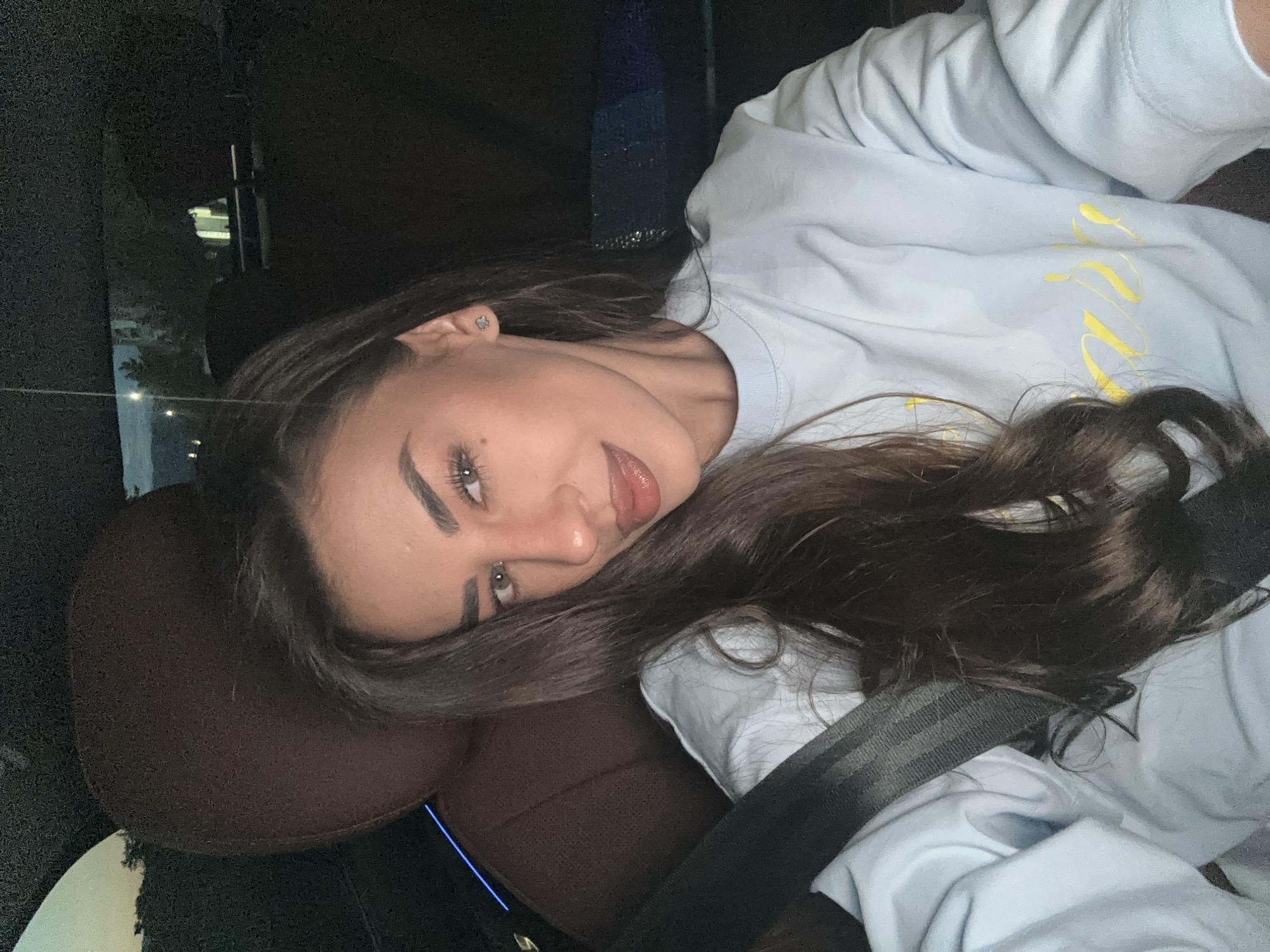 A woman is taking a selfie in a car at night. She is sitting in the driver's seat, and a seatbelt is fastened across her chest. She has long, dark hair and is wearing a light blue sweatshirt with yellow lettering. The interior of the car is dark, with the exception of some ambient lighting visible on the car seat and through the window. Outside the car, city lights and trees are visible in the distance. Based on the available information, the location of this scene is the city of Petrich, Bulgaria.