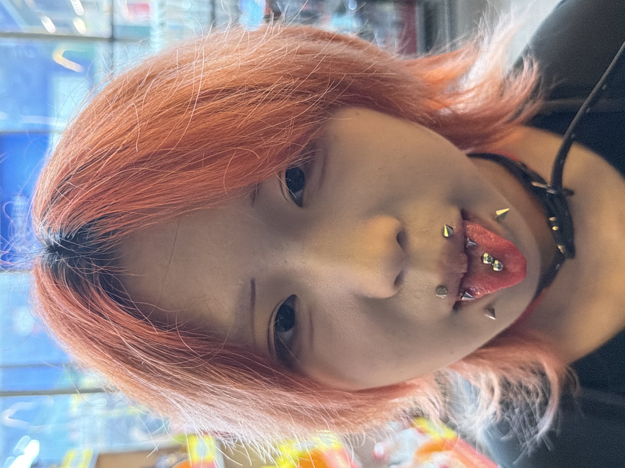 A young individual with orange-dyed hair and dark roots gazes directly forward, their tongue protruding. The person has multiple facial piercings: two spike-stud piercings on the upper lip, two spike-stud piercings on the lower lip, and two spherical-ended barbell piercings on the tongue. Additionally, two spike-shaped dermal or surface piercings are visible on each cheek. A red choker is worn around the neck, underneath a black choker or harness-style accessory adorned with spike studs. The individual wears a dark-colored top. The background is an indistinct, highly blurred indoor environment, characterized by soft, bright, multicolored bokeh lights, consistent with a retail or commercial setting. The specific location within 江北城街道, China, is not visually ascertainable from the image.
