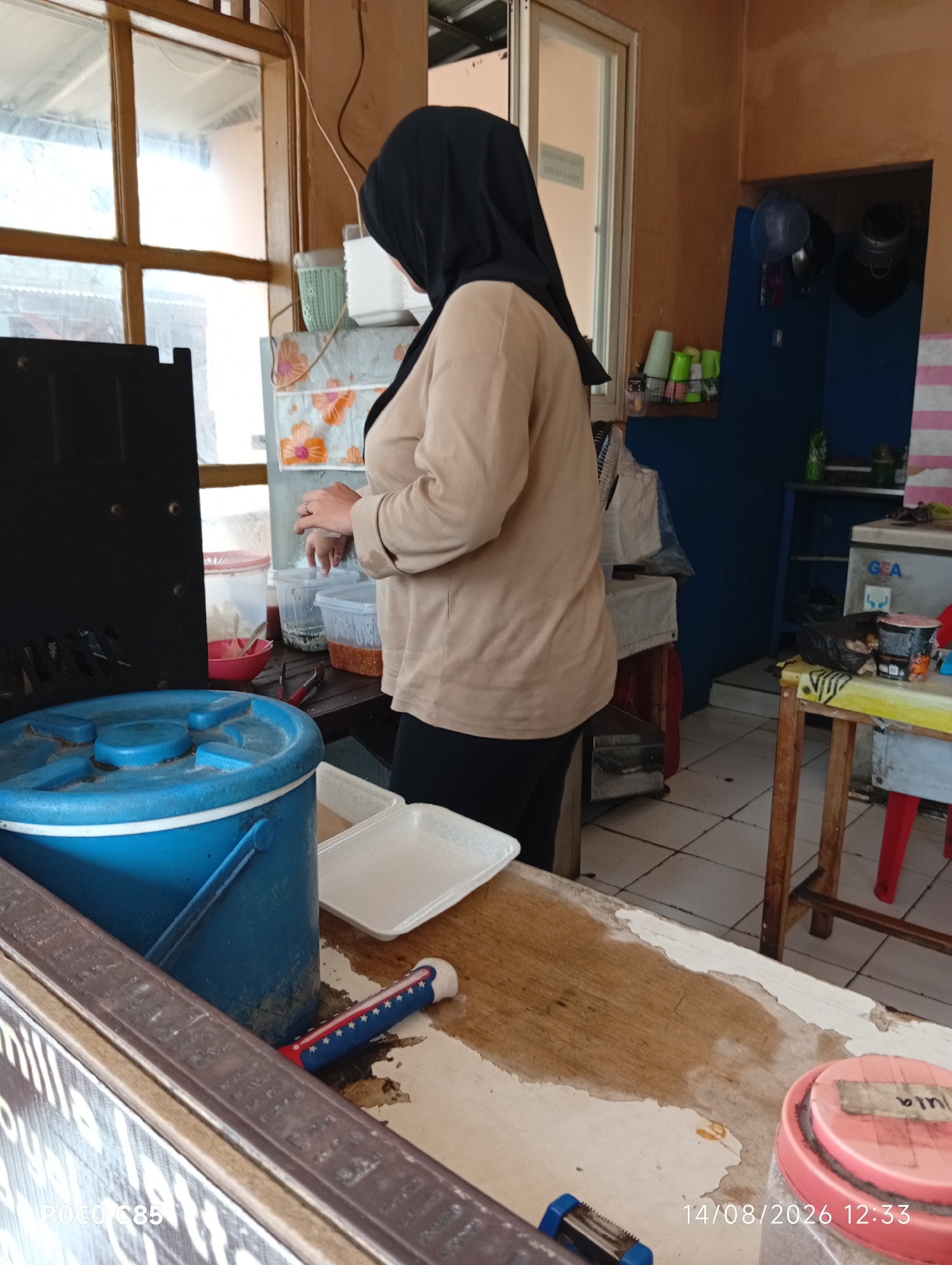 In a seemingly open-air or semi-enclosed establishment, possibly a food stall or small eatery in South Tangerang, Indonesia, a person wearing a black hijab and a beige long-sleeved shirt stands behind a wooden counter. The person's hands are visible, with one hand resting on the counter and the other seemingly in the process of handling an object.
The counter is cluttered with various items, including a large blue bucket, white disposable food containers, and a red bowl with a red object inside. To the left of the counter, a black appliance or piece of equipment is partially visible. In the background, shelves are stocked with items, including bottles and containers. A blue-tiled wall is visible behind the counter, with a small fan mounted on it.
A window with wooden frames is present on the left side of the frame, offering a view of an outdoor area. The flooring appears to be tiled. A portion of a sign with text, possibly bearing the brand "POCO C85", is visible at the bottom left. A date stamp, "14/08/2026 12:33", is present in the bottom right corner of the image. The overall impression is one of a functional workspace, possibly involved in food preparation or service.