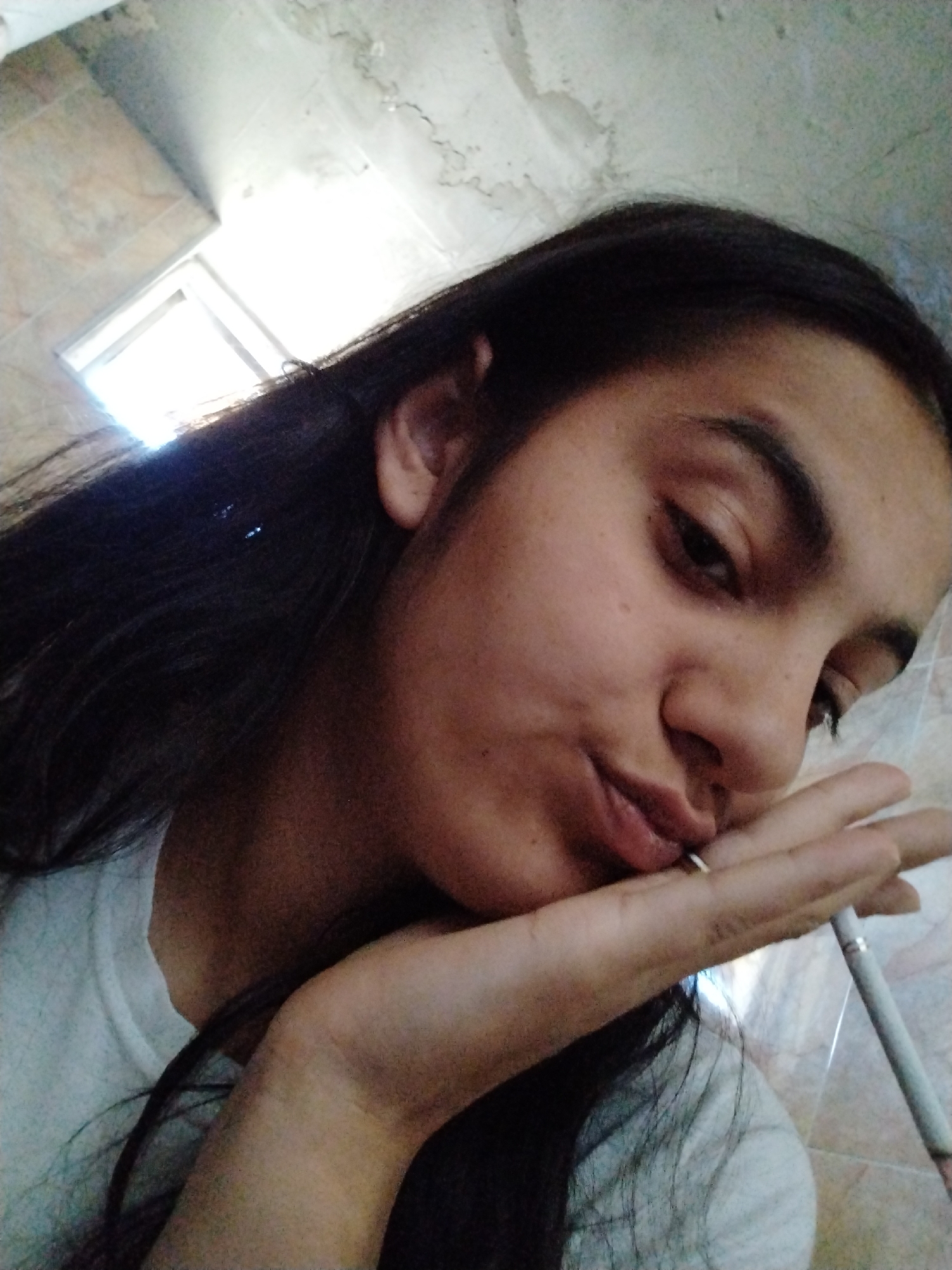A woman is indoors holding a cigarette to her lips. Her long dark hair falls around her face, and she is wearing a white shirt. The location appears to be a bathroom, as suggested by the wall tile visible in the background and the light fixture above. The city and location are Unknown, Unknown.