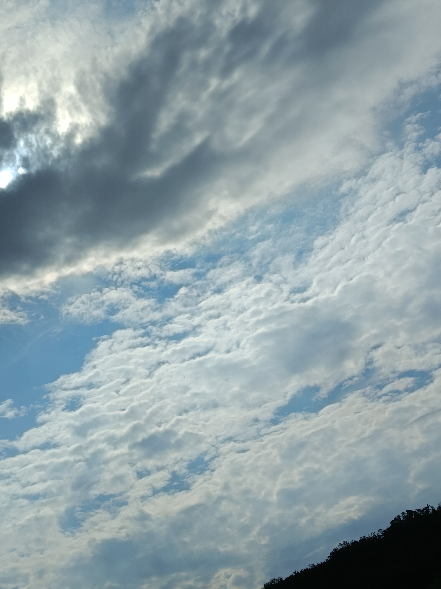 The image shows a cloudy sky above a silhouetted treeline. The sun is partially obscured by dark clouds in the upper left quadrant. Lighter, wispy clouds fill the rest of the sky, with patches of blue visible. The treeline forms a dark, uneven band across the bottom of the frame, suggesting a hilly or mountainous terrain. The scene appears to be a natural landscape. The image does not contain any information or visual cues that would allow for its placement in the city of Razeni, Moldova.