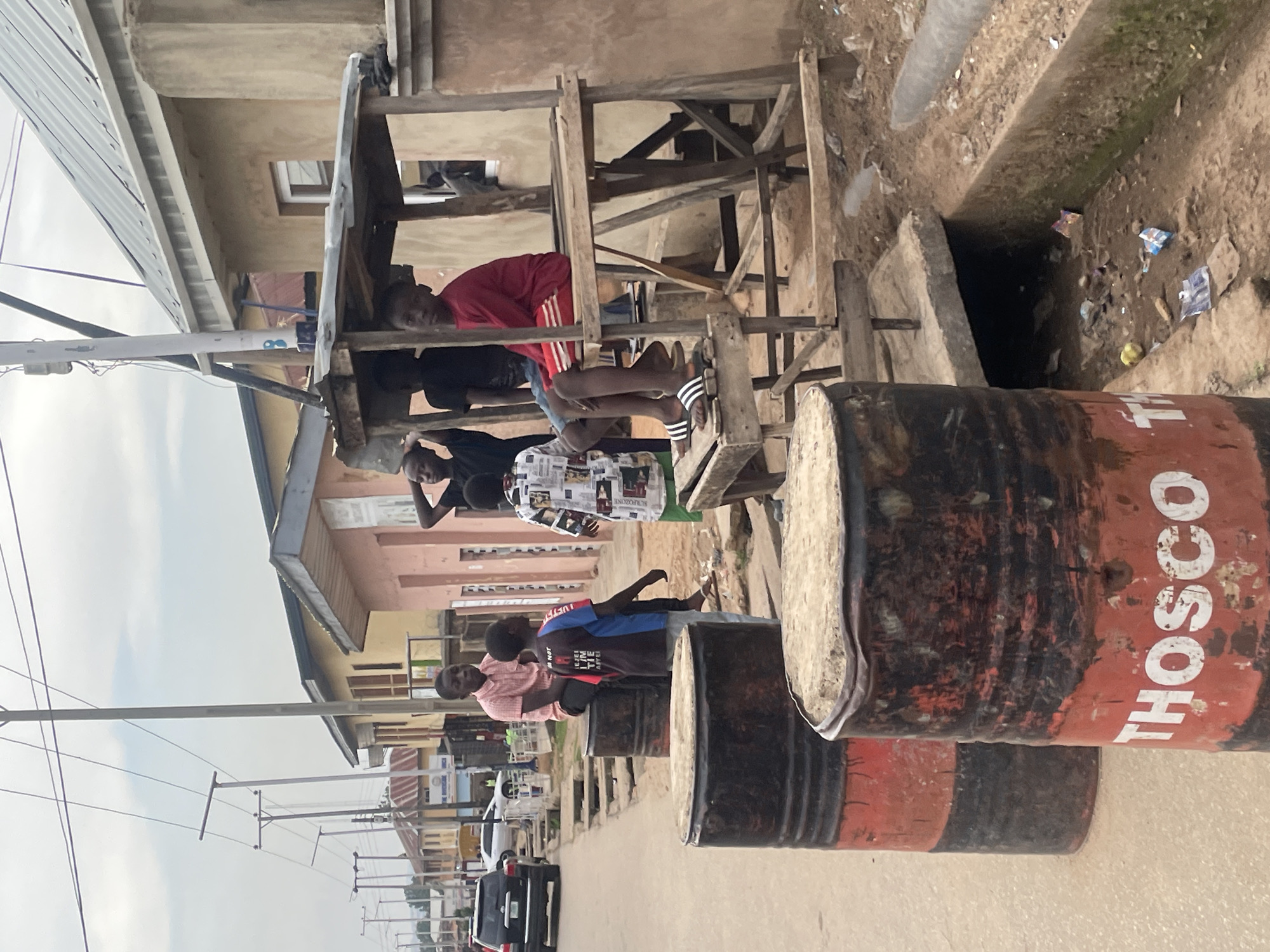 The image depicts a street scene in Jos, Nigeria. Several young men are positioned on and around a makeshift wooden structure built over a drainage ditch. The structure is constructed with crude lumber and appears unstable. A row of oil drums, some with the word "THOSCO" painted on them, line the roadside. The men appear to be loitering or socializing. The buildings in the background are a mix of residential and commercial structures, indicating a neighborhood setting. A street with cars can be seen in the distance. The overall impression is a scene of informal gathering in a somewhat impoverished urban environment.