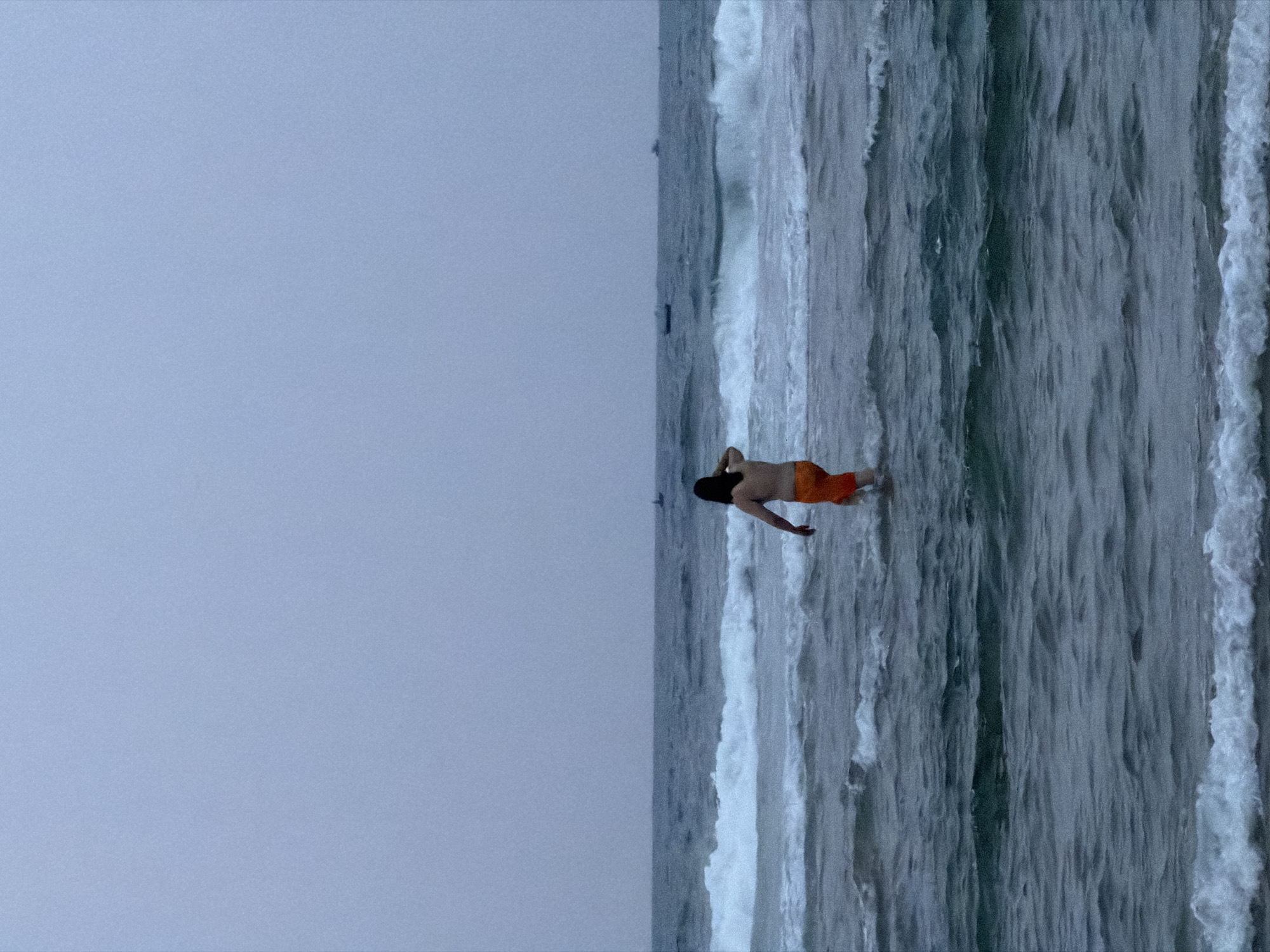 The image shows a solitary person wading in the ocean under an overcast sky. The horizon line runs horizontally across the middle of the frame, dividing the muted grey-blue sky from the active sea. An adult male, viewed from behind, stands in the water, which reaches approximately his waist. He has dark hair, a bare upper body, and is wearing bright orange swim shorts. His arms are slightly bent, and his posture suggests he is either entering the water, standing, or preparing to move further.
The ocean exhibits dynamic conditions with multiple lines of white-capped waves breaking and rolling towards the shore. The water appears dark and deep, reflecting the overcast light. The sky is uniformly light blue-grey, indicating a cloudy or hazy day. In the far distance, on the horizon, a faint, dark shape might suggest a distant boat or a small landmass. The scene evokes a sense of calm and contemplation, with the lone figure immersed in the vastness of the sea and sky. The overall atmosphere is quiet and natural. No visible text is present in the image. The location is consistent with a coastal area in Đà Nẵng, Vietnam.