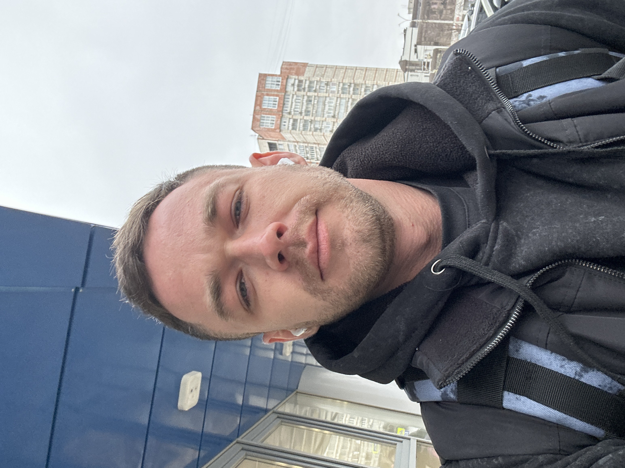 A man with fair skin, short blonde hair, and a stubbly beard is pictured in an outdoor selfie. He is wearing a black hooded jacket with black and blue striped detailing and has white wireless earbuds in both ears. The man is positioned near a building with blue paneling and glass windows, likely a commercial structure. In the background, a tall, multi-story building with a brick facade and rows of windows can be seen under an overcast sky. Given the information about the city, the location can be identified as Perm, Russia.