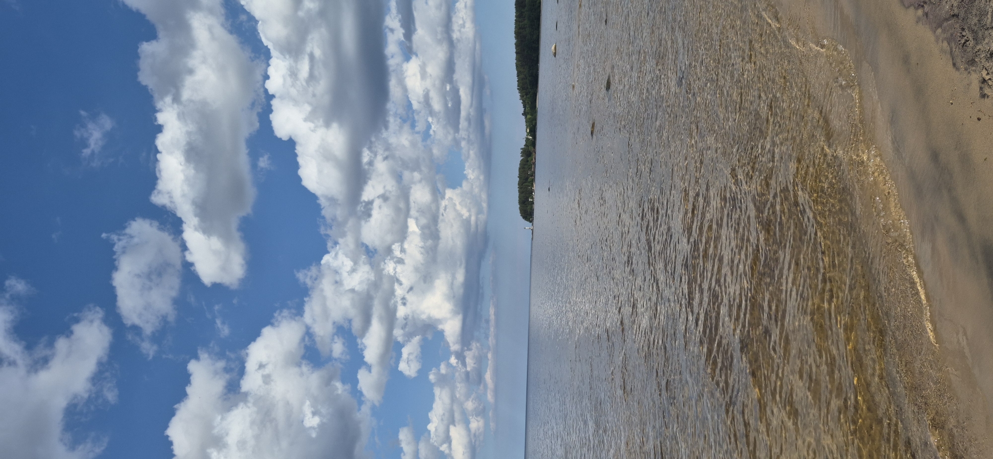 The image depicts a natural landscape of a lake or sea under a partly cloudy sky. The foreground shows a sandy shore with shallow, clear water lapping at the edge. The water is slightly murky, revealing the sandy bottom. In the mid-ground, the water stretches out to the horizon, meeting a distant tree-lined shore. On this distant shore, a white lighthouse is visible to the left, and a line of trees forms a dense green band against the sky. The sky is a vibrant blue with scattered white and gray cumulus clouds, some of which cast shadows on the water. The overall scene conveys a sense of tranquility and natural beauty. The location is identified as Motornoe, Russia.