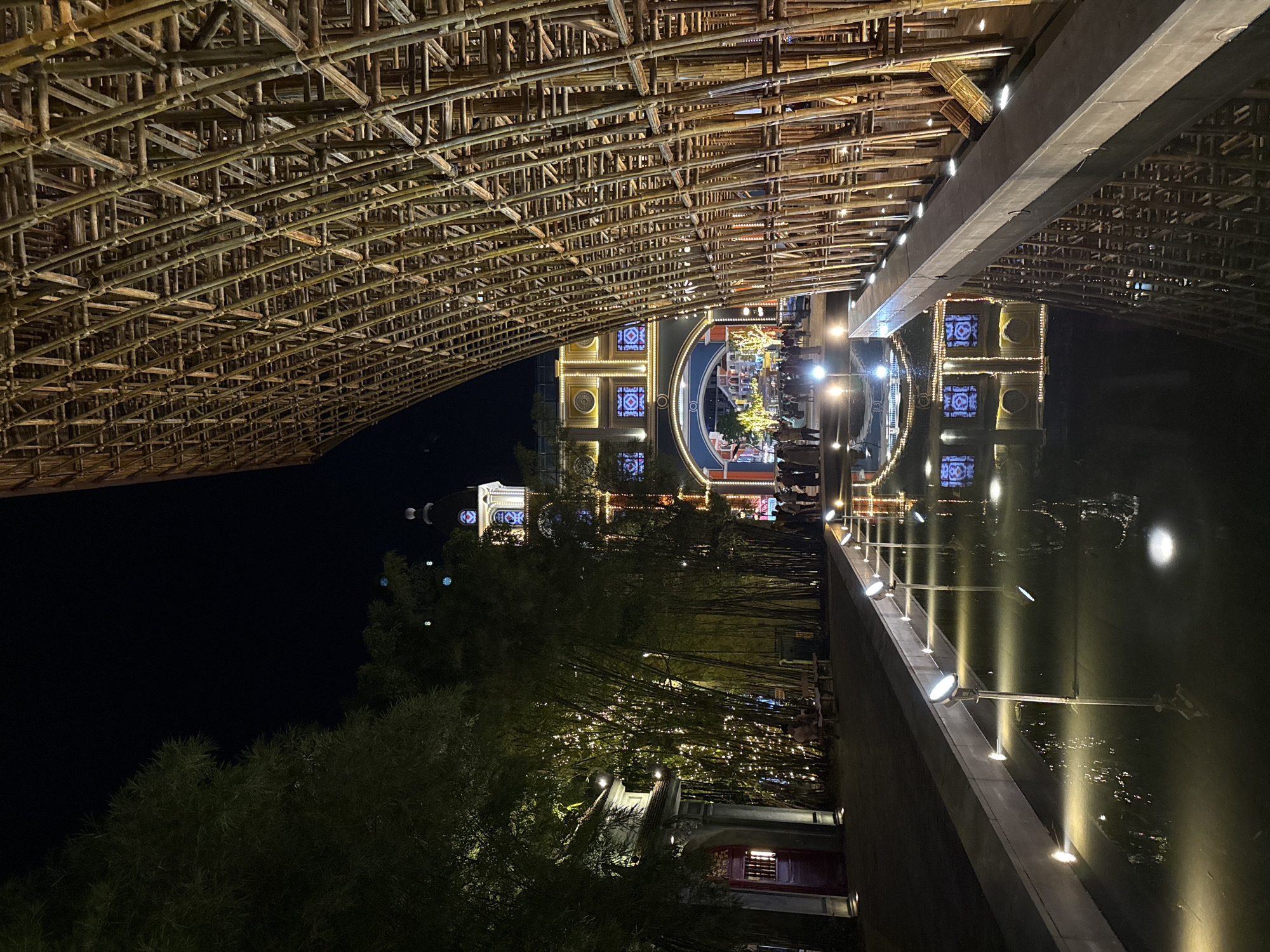 This image depicts a vibrant night scene at a beautifully lit tourist attraction in Phú Quốc, Vietnam. The most prominent feature is a massive, intricately woven structure made of bamboo poles, forming a curved overhead canopy or bridge. This bamboo architecture is illuminated from below, highlighting its complex, gridded patterns and natural texture against the dark night sky.
Below this impressive structure, a calm canal or waterway reflects the numerous artificial lights. The near bank features a concrete walkway or low wall, equipped with a series of spotlights that cast bright, distinct beams onto the water and the opposite side.
Along the far bank of the canal, several ornate buildings are brightly lit, showcasing architectural details such as arches, circular windows, and decorative motifs. One central building stands out with a large, glowing circular archway. Interspersed among the buildings and lining the banks are lush bamboo trees and other dense greenery, with some trees adorned with string lights, enhancing the festive atmosphere.
Small groups of people can be seen strolling or gathered on the far bank, suggesting a popular evening destination for visitors. The scene conveys a lively and enchanting ambiance, characteristic of a well-designed and illuminated outdoor attraction after dusk. The sky is dark, indicating nighttime conditions, and there is no discernible text visible in the image.