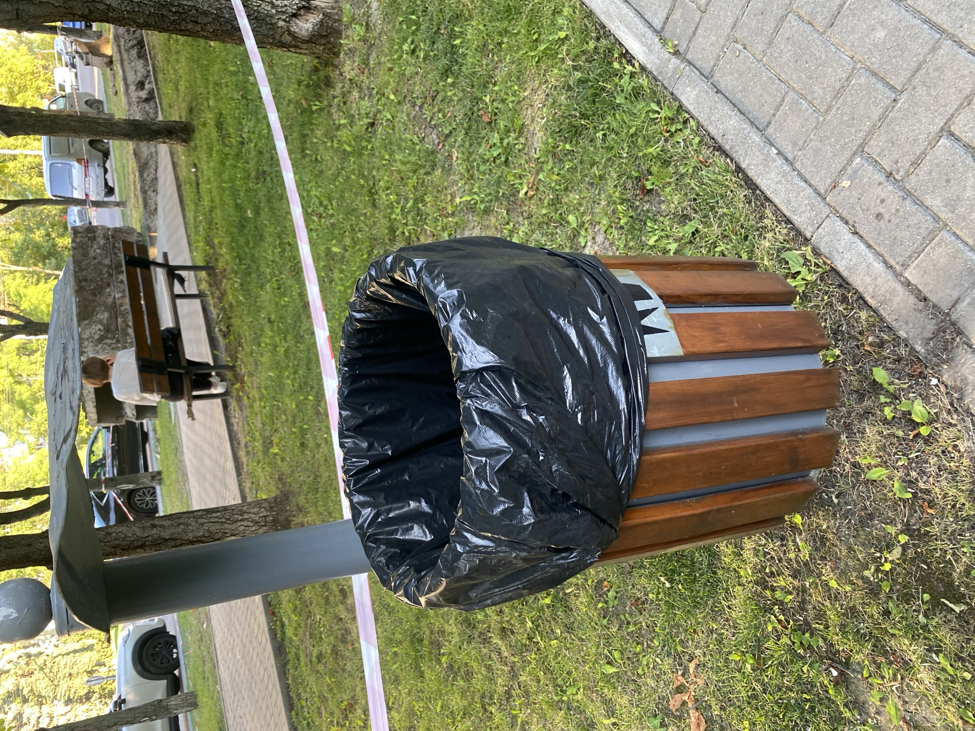 A trash can with a black plastic liner stands on a paved sidewalk next to a grassy area. In the background, a person sits on a park bench facing away from the camera. Beyond the bench, a street is visible with cars and construction activity. A pink and white caution tape is draped across the scene. The image appears to be taken in a public park or street in Chișinău, Moldova.