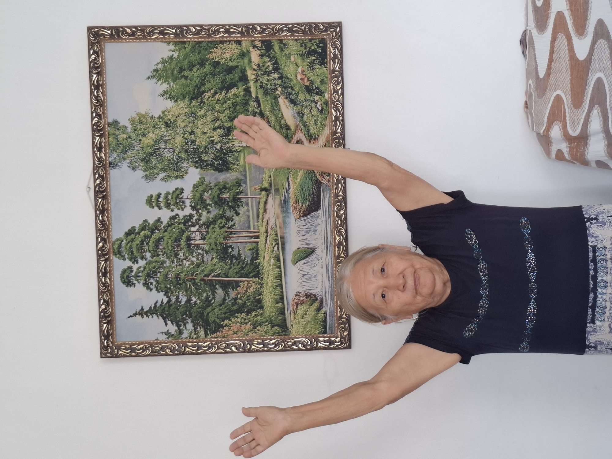An elderly woman with gray hair pulled back stands in front of a painting of a forest with a waterfall. She is wearing a dark blue t-shirt with a patterned design and a patterned skirt. Her arms are raised, and her palms are open and facing forward. She is looking directly at the camera with a neutral expression. The painting is framed in an ornate gold frame and hangs on a white wall. To the right of the woman, a portion of a sofa with a patterned throw is visible. The lighting in the image suggests an indoor setting, possibly a home. There are no clear indicators of the specific location within Almaty, Kazakhstan, beyond it being an indoor environment.