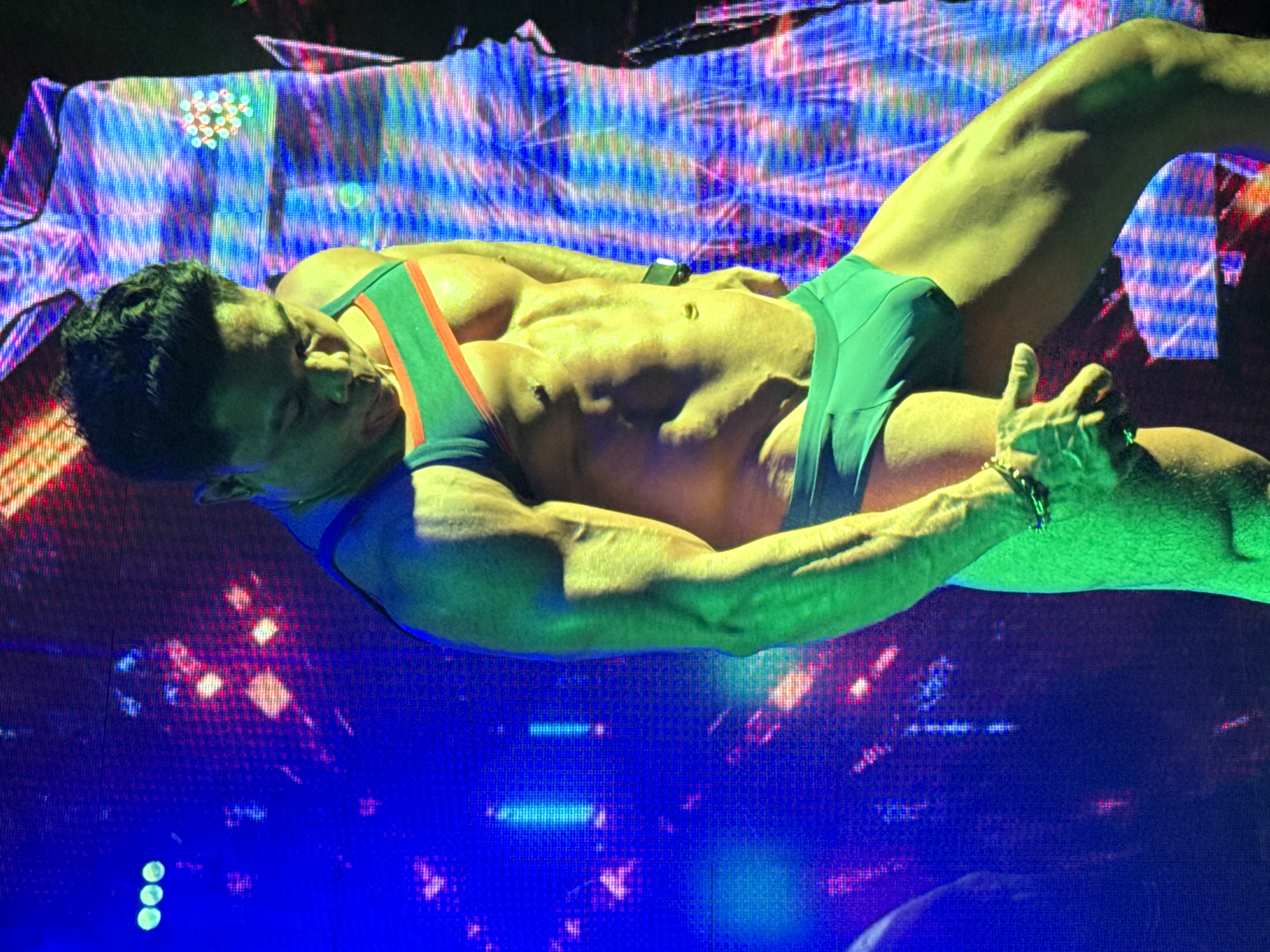 A muscular man in a green singlet with a red stripe lies on his back against a backdrop of blue and purple lights. He is positioned horizontally with his head to the left and his legs angled upwards to the right. His eyes are closed and his mouth is slightly open. The man is wearing a bracelet on his left wrist. The background consists of blurred, colorful lights arranged in a digital-appearing pattern. There is no information to identify this location as Kuta, Indonesia from the image.