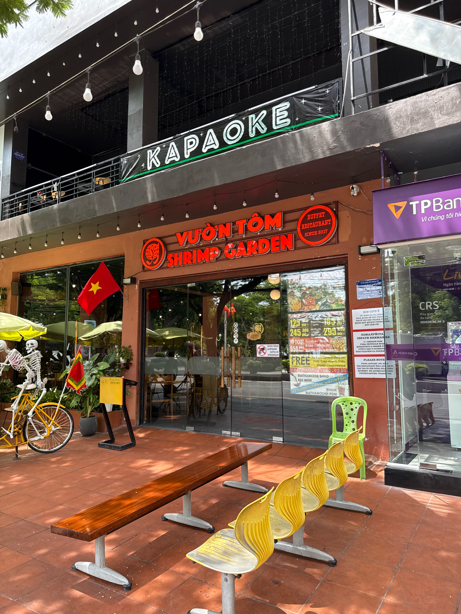 The image captures a street-level view of a restaurant called "VUON TOM SHRIMP GARDEN" in Nha Trang, Vietnam. The restaurant's prominent orange signage with red lettering is clearly visible above the glass-fronted entrance. To the left of the entrance, a yellow bicycle with a skeleton rider and two flags are displayed. Outside the restaurant, there are several outdoor seating options, including a long wooden bench and a row of yellow chairs. A bright green polka-dotted chair is positioned near the entrance. In the background, above the restaurant, a banner advertising "KARAOKE" is displayed. A TPBank ATM is situated to the right of the restaurant. The ground is paved with reddish-brown tiles, and trees are visible in the upper left corner of the image. The scene appears to be during daylight hours, with sunlight casting shadows on the ground.