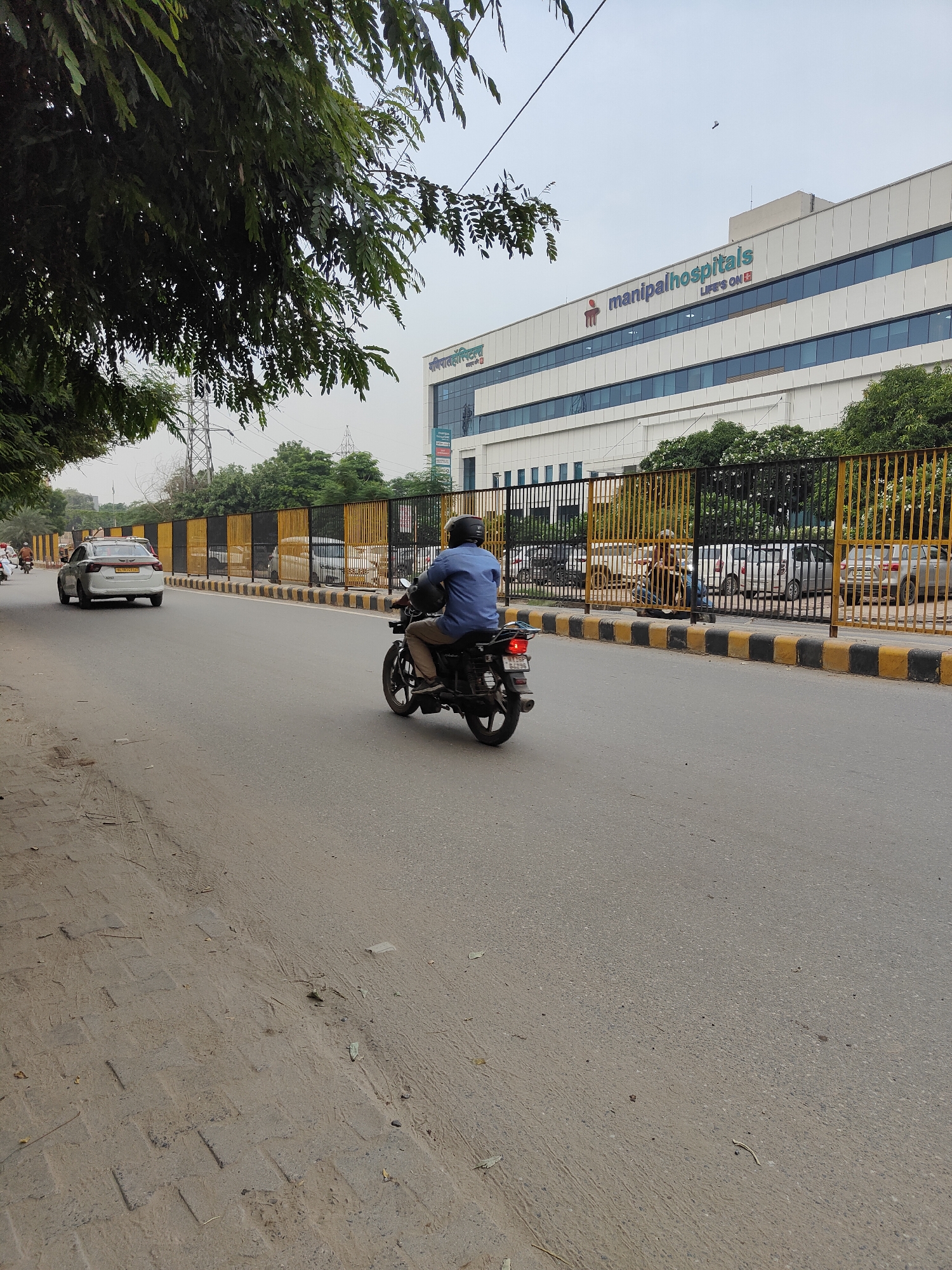 The image shows a street scene in Gurgaon, India, with a white sedan driving in the left lane and a motorcyclist in the right lane. In the background, a large modern building with "manipalhospitals" signage is visible. A black and yellow striped fence separates the road from the hospital property. Trees and electrical poles are also present. The sky is overcast.