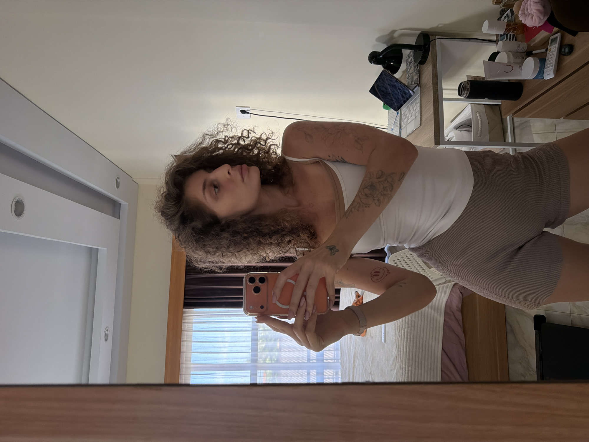 A woman with curly brown hair, wearing a white tank top and brown shorts, is taking a mirror selfie in what appears to be a bedroom. She is holding a pink smartphone with three camera lenses. The room has a bed with white and pink bedding, a window with sheer curtains, and a wooden desk with a laptop, a lamp, and various cosmetic products. There are tattoos visible on her arms, including a smiley face with melting features and a butterfly. The location is identified as North Kuta, Indonesia.