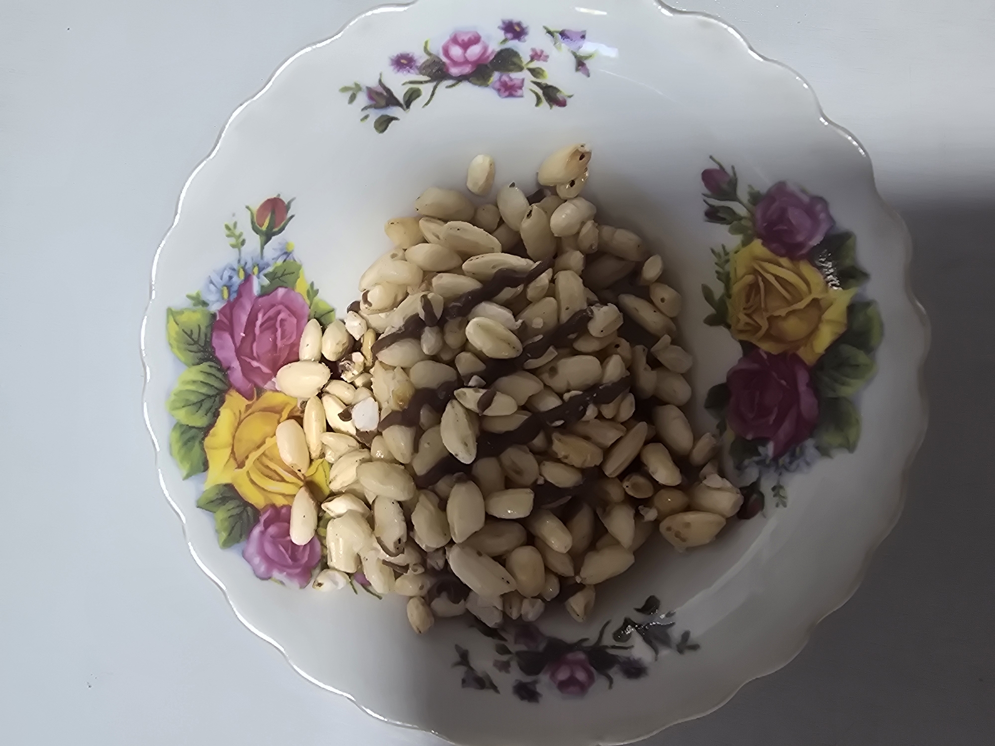A top-down, medium shot captures a white porcelain plate adorned with a floral pattern featuring roses in yellow, pink, and purple, along with green leaves and small blue flowers. The plate is filled with blanched almonds, some of which appear to be drizzled with dark chocolate. The almonds are piled in the center of the plate, obscuring part of the floral design. The background is a plain, light-colored surface, possibly a table. There is no identifiable location in the city of Ulan-Ude, Russia, and no people or actions are depicted.