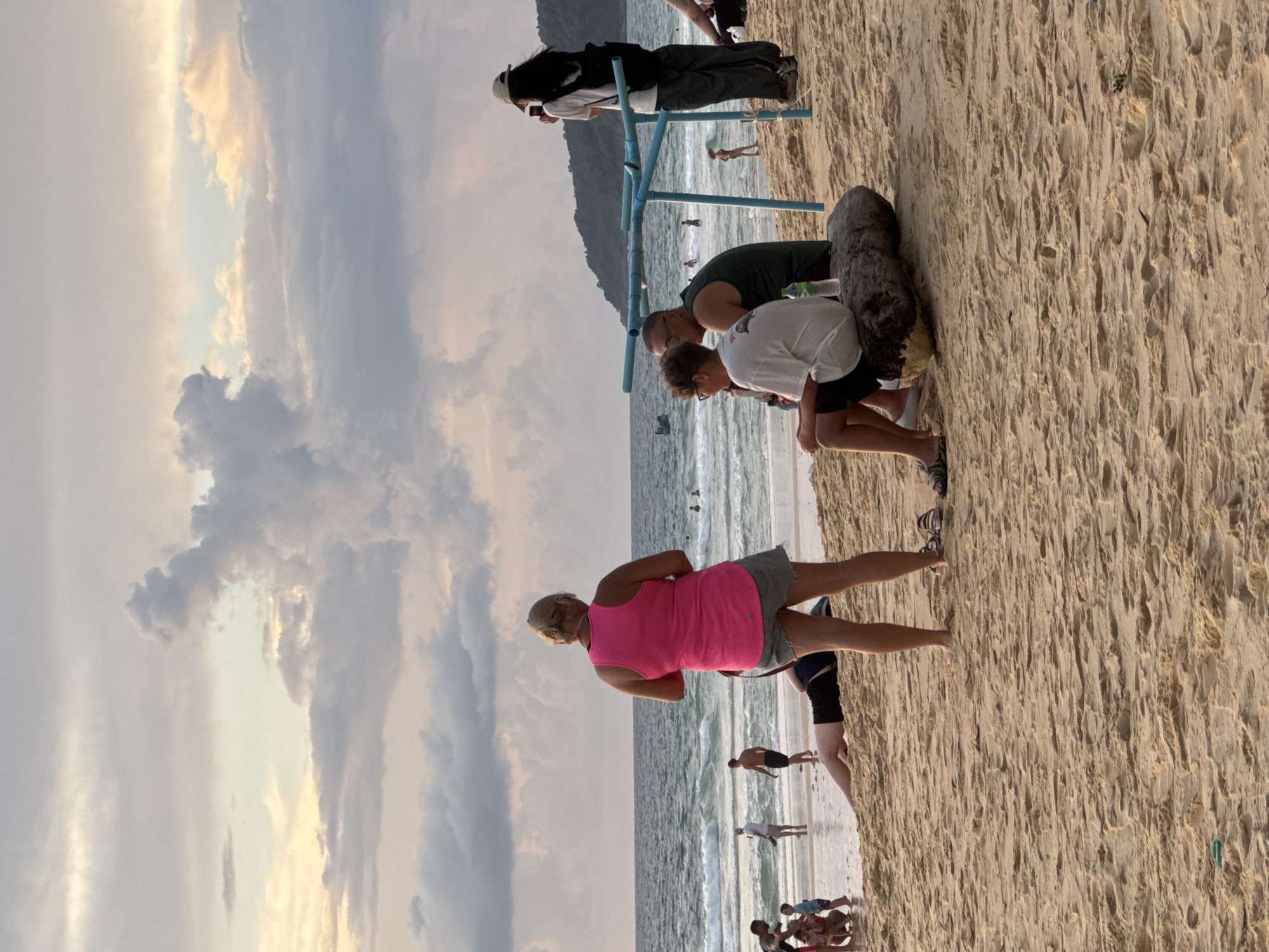 A group of people are on a sandy beach in Karon, Thailand, with the ocean and a cloudy sky in the background. In the foreground, a woman wearing a bright pink tank top and gray shorts stands facing away from the camera, looking towards the sea. To her right, three men are seated on a large piece of driftwood. One man wears a white t-shirt and black shorts, another wears a dark green tank top, and the third man, seen from the side, wears a white t-shirt and dark shorts. To the right of these men, a woman with long dark hair, wearing a white t-shirt, dark pants, and a beige hat, is taking a picture with her phone.
In the mid-ground, several other individuals are visible near the water's edge. Some are walking in the shallow surf, while others are further out in the sea. A person is lying down on the sand in the foreground, to the left of the seated men. The beach is covered in fine, light-colored sand, with some footprints and scattered debris. The ocean has moderate waves, and a few small boats are visible in the distance. The sky is filled with a mix of dark and lighter clouds, with hints of pale yellow and pink light suggesting it may be near sunset. A hilly, tree-covered landscape is visible on the horizon to the right.