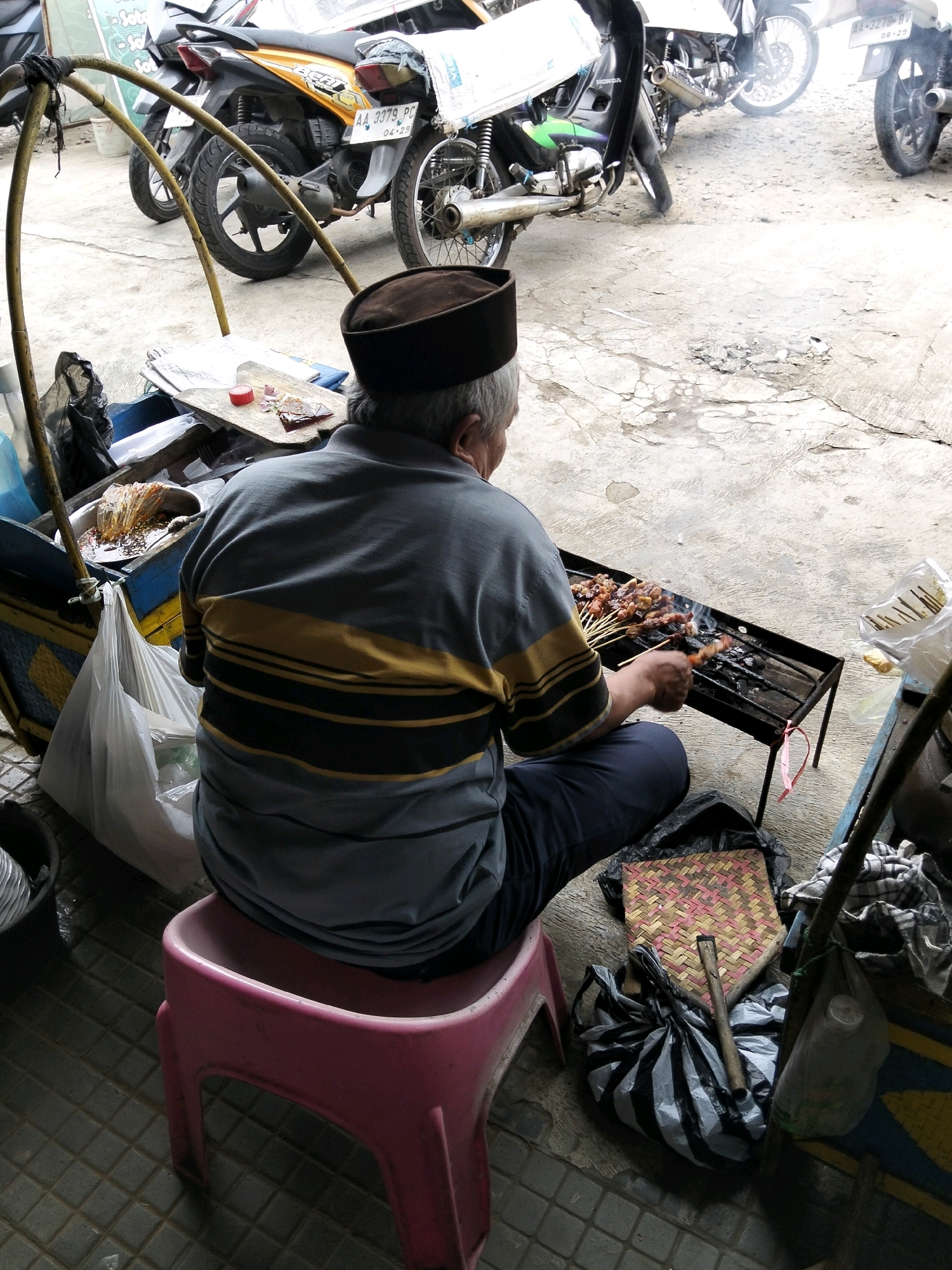In Kemiri Kidul, Indonesia, a man wearing a brown cap and a grey shirt with yellow and black stripes sits on a pink plastic stool, grilling satay over a charcoal brazier. The scene appears to be outdoors, possibly at a street-side stall, with motorcycles parked in the background. The ground is concrete, and various items, including plastic bags, a woven mat, and a container of satay sauce, are scattered around the area. The man is actively tending to the satay, using a stick to turn the skewers over the hot coals. Smoke rises from the grill, indicating the cooking process is underway. The overall impression is that of a food vendor preparing to serve customers.