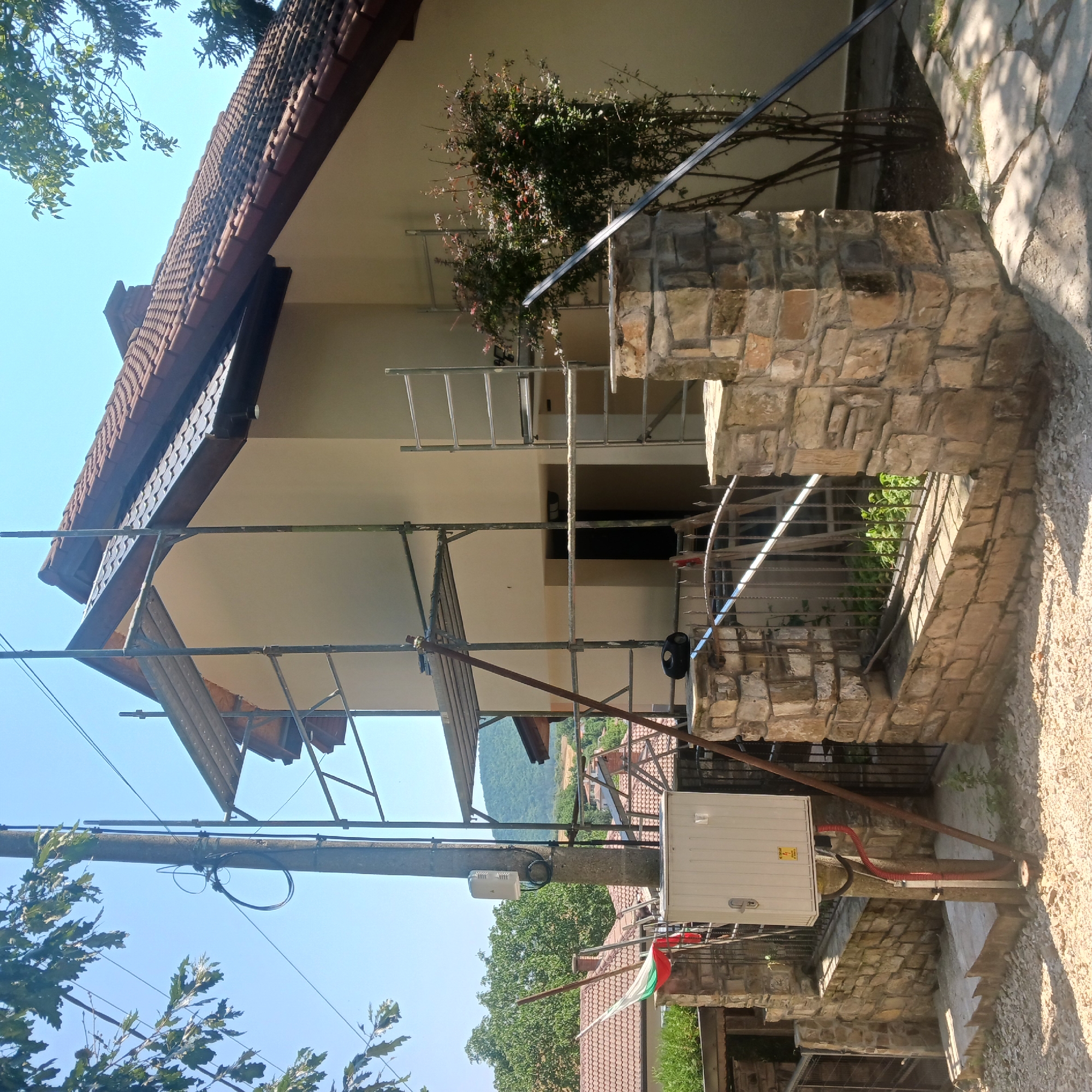 The image depicts a house undergoing renovation or construction, as evidenced by the scaffolding surrounding a portion of the structure. The house appears to be located in a rural or suburban area, with trees and rolling hills visible in the background. A Bulgarian flag is visible, partially obscured by a metal fence and a stone wall. A utility pole with electrical boxes and wiring is situated to the left of the house. The overall scene suggests an ongoing project in a residential setting. The location is confirmed to be Sulitsa, Bulgaria.