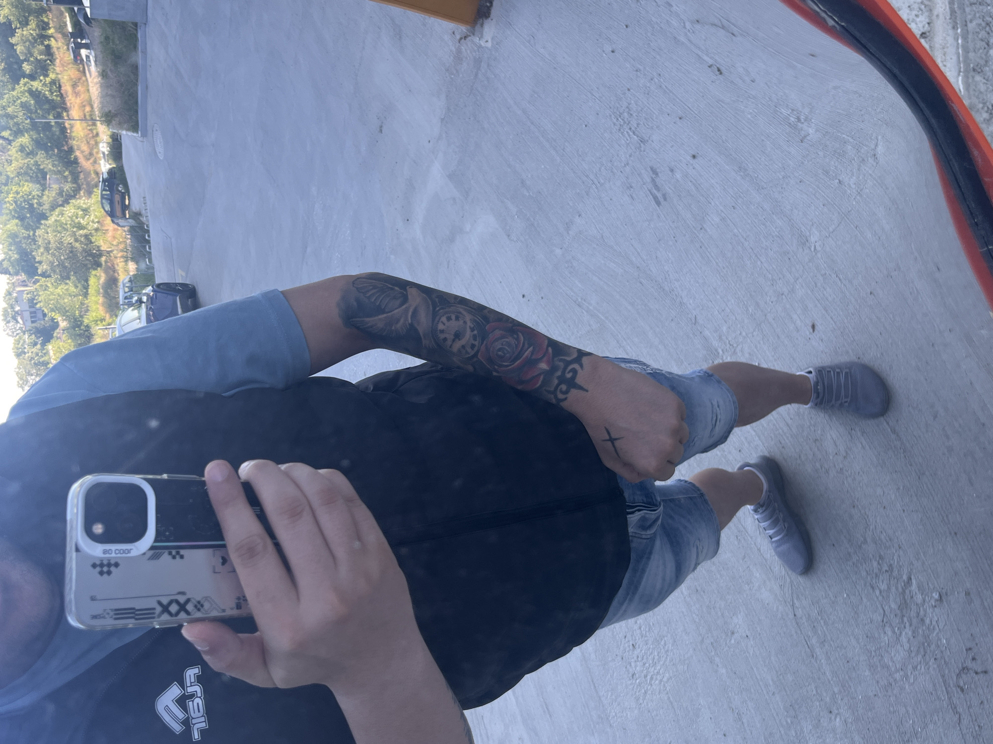 A person, viewed from the waist down, stands on a concrete surface. They are wearing denim shorts, a blue t-shirt, and a black vest. Grey sneakers are on their feet. On their left arm, there is a tattoo featuring a dove, a clock, and a rose. Their right hand is clenched into a fist, with a small cross tattoo visible on the back of it. The person is holding a smartphone in their left hand, which is angled to capture a reflection of themselves and the surrounding environment.
In the background, a paved incline leads uphill. A dark-colored car is parked on the side of the road further up the incline. A blue SUV is visible on the left side of the road, also on the incline. Trees and vegetation line the right side of the incline, with some buildings visible through the trees at the top. The lighting suggests it is daytime, with shadows cast on the concrete. The overall environment appears to be an outdoor urban or suburban setting, potentially a parking lot or a street in Varna, Bulgaria, given the typical architecture and landscape observed in such locations.