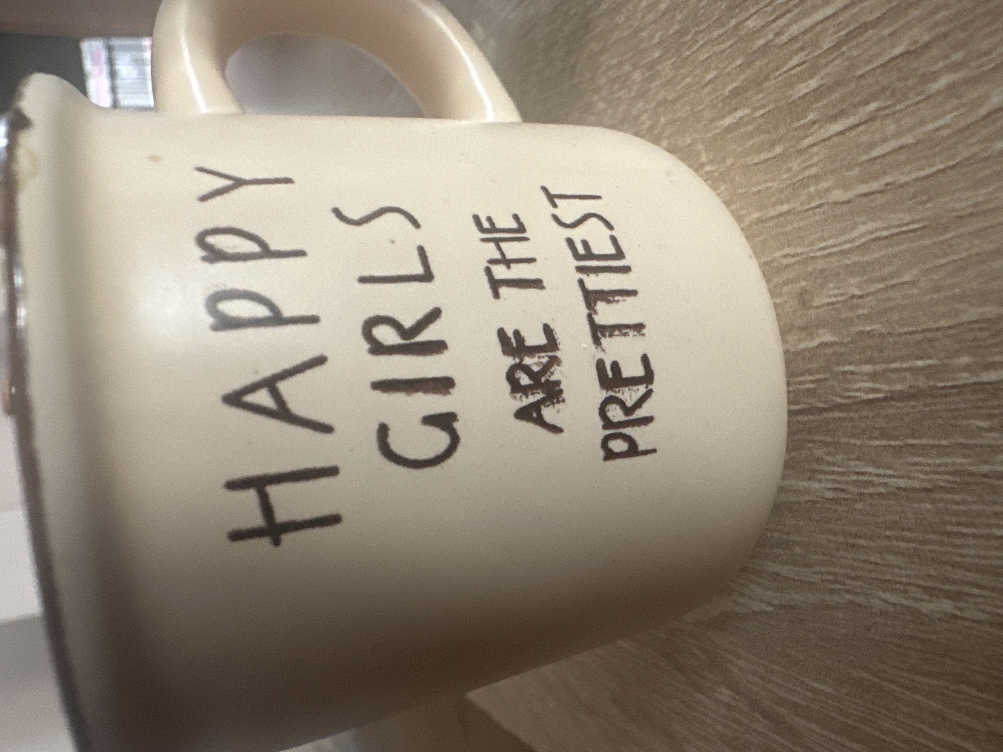 A close-up shot shows a cream-colored mug with the phrase "HAPPY GIRLS ARE THE PRETTIEST" written in black lettering. The mug is sitting on a surface with a wood-grain pattern. No people or specific environmental context related to Sofia, Bulgaria, are visible in the image.