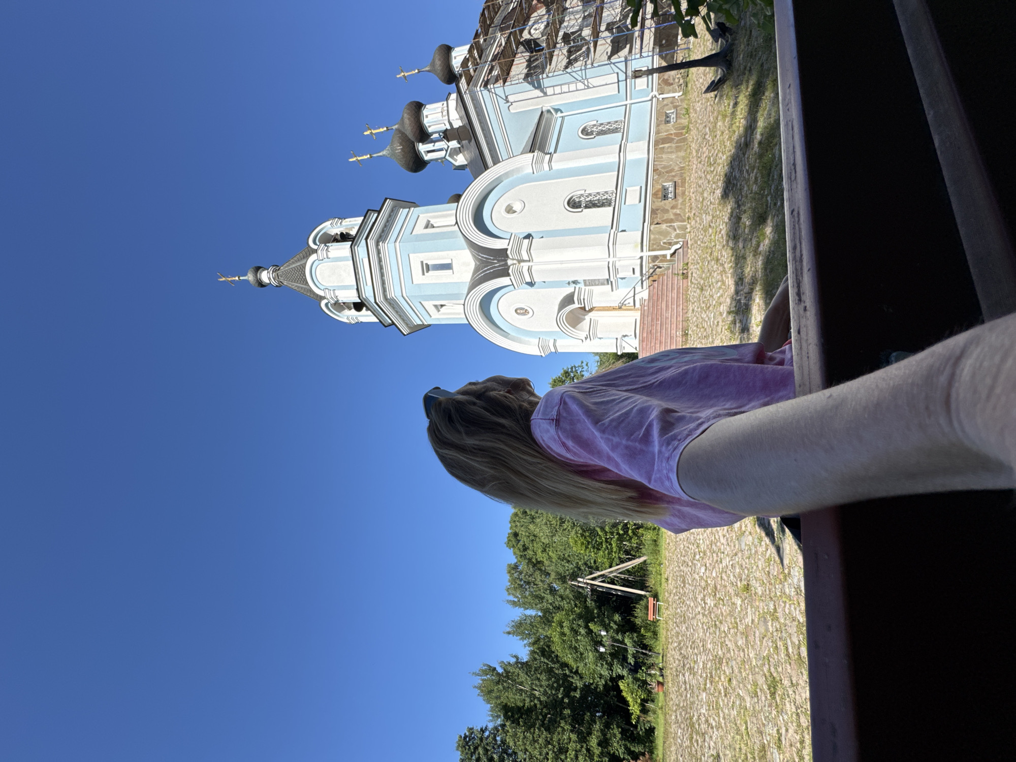 A woman with long blonde hair, wearing a pink tie-dye t-shirt and sunglasses, is standing in front of a light blue and white Orthodox church with multiple domes, located in St. Ozerki, Russia. The church appears to be under renovation, with scaffolding visible on one side. The ground in front of the church is paved with stones, and there are trees and green grass in the background. The sky is clear and blue. The photo is taken from a low angle, with the woman's arm and hand in the foreground, suggesting a selfie or a photo taken by someone close to her.