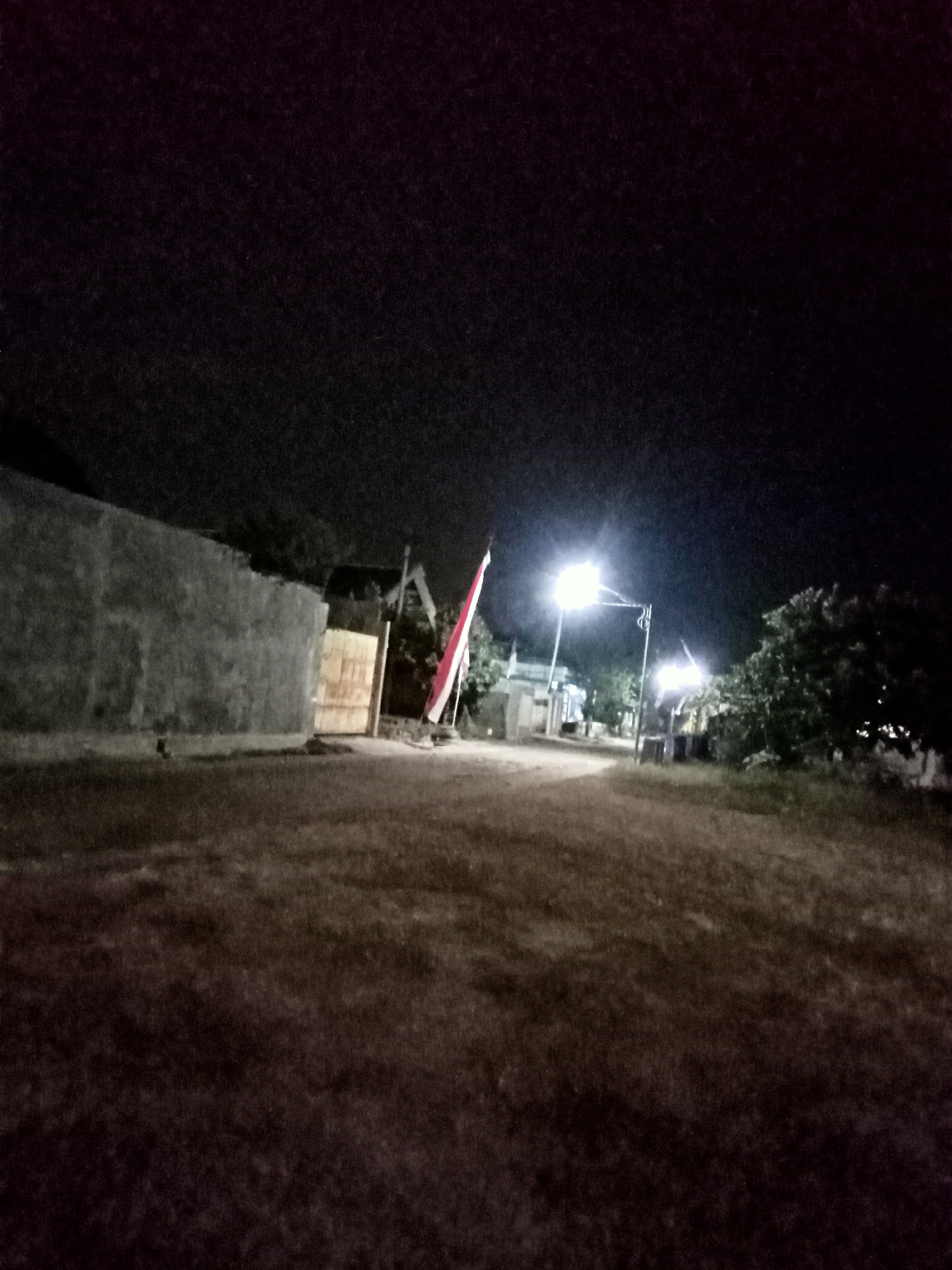 The image depicts a dimly lit street scene at night in the city of Simo, Indonesia. A rough, textured concrete wall dominates the left side of the frame, extending into the background. Adjacent to the wall is a light-colored wooden gate or fence. To the right of the gate, a red and white flag, likely the Indonesian flag, is prominently displayed, attached to a pole. In the background, streetlights illuminate the path, casting a glow on the surrounding area. Trees and simple buildings are visible in the distance, their details obscured by the darkness. The ground in the foreground is covered with dry grass or soil, appearing uneven and sparsely vegetated. The overall impression is of a quiet, residential street at night, with a national symbol indicating a sense of place and identity.