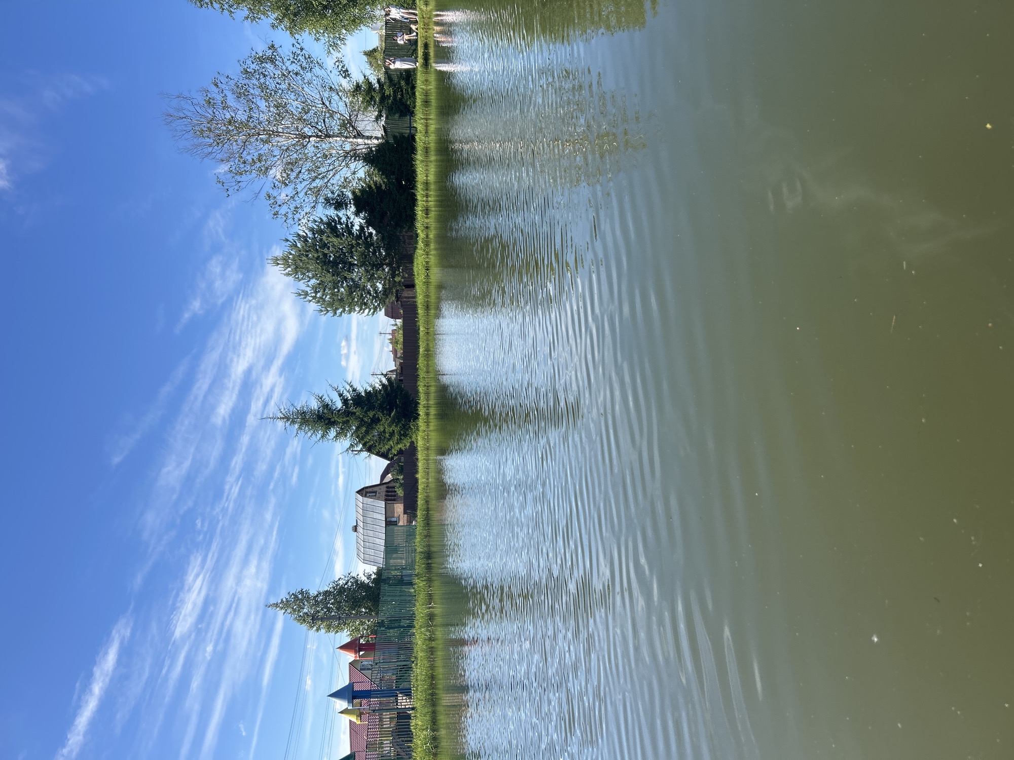The media file depicts a serene waterscape under a bright blue sky with wispy white clouds. The scene is a reflection of a green, grassy bank lined with trees and several small buildings. A wooden fence runs along the edge of the bank. In the foreground, the calm water exhibits gentle ripples, reflecting the sky and the terrestrial elements. The water appears slightly murky and a pale green color.
On the bank, several structures are visible, including what looks like a greenhouse or a shed with a metal roof, and a more traditional building with a brick facade and a gabled roof. Behind these, more trees can be seen, some evergreen and others deciduous. To the left of the frame, a children's playground with colorful structures is partially visible. In the far distance, on the water's surface, faint reflections suggest the presence of people, though they are indistinct.
The lighting suggests it is daytime, likely a sunny day given the clear blue sky and bright reflections on the water. The overall atmosphere is peaceful and tranquil. The location context, "Слава Снт, Russia," indicates the setting is likely a garden community or dacha area in Russia. There is no visible text in the image. The image is oriented sideways.