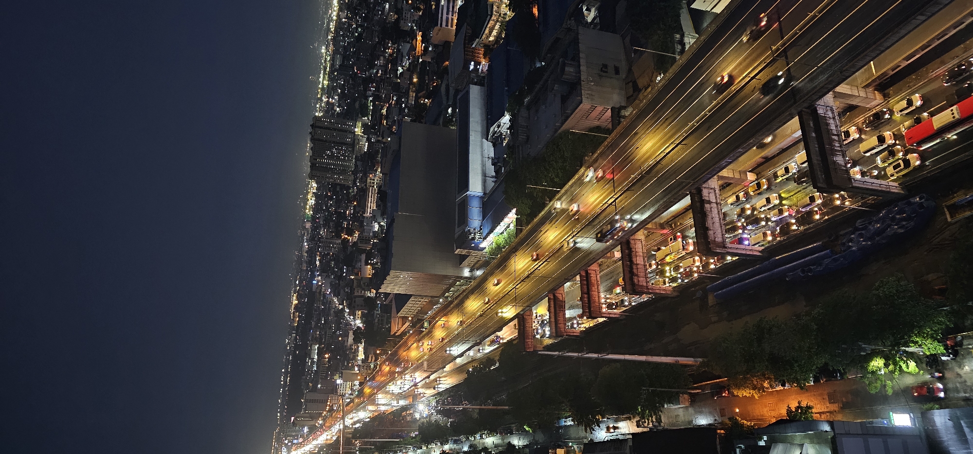 The image captures an aerial view of a busy highway at night, likely within the city of Makati, Philippines, characterized by its dense urban landscape. A multi-lane elevated highway dominates the foreground, illuminated by the headlights and taillights of numerous vehicles. The highway appears to be either under construction or undergoing maintenance, with visible scaffolding and barriers along its edges. Traffic is heavy, with cars, buses, and trucks forming long queues, their lights creating streaks of color against the dark asphalt.
Below the elevated highway, another road or underpass is visible, also congested with vehicles. The surrounding area is a dense cityscape, with buildings of varying heights and architectural styles lit up, suggesting a bustling urban environment. The sky is dark, typical of nighttime, with no discernible celestial bodies. The overall scene depicts a lively, continuous flow of traffic and urban activity, even in the late hours, highlighting the constant movement and energy of a major metropolitan area. The presence of lights from buildings and vehicles indicates a well-developed and populated urban center.