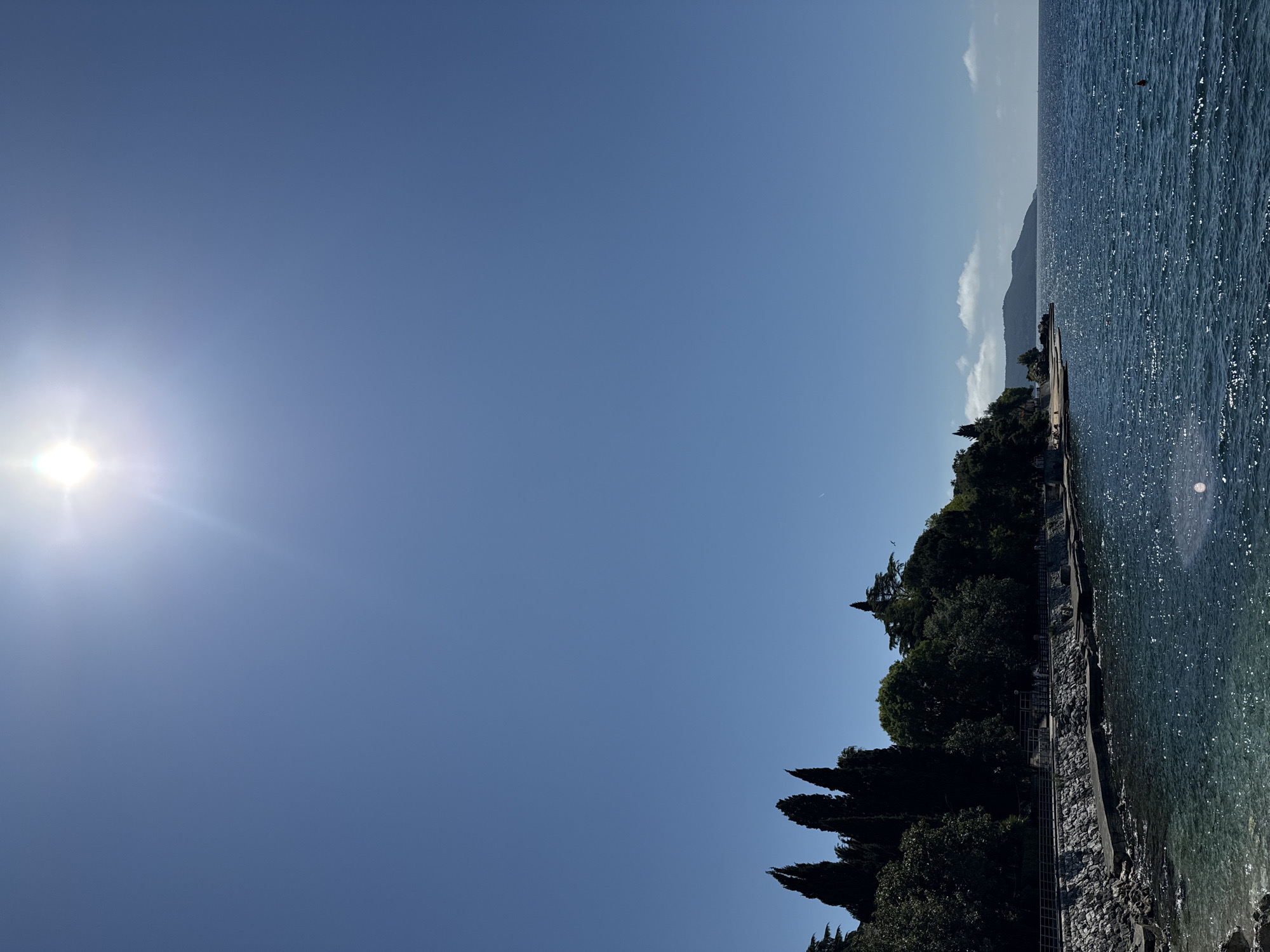 The image depicts a coastline scene under a clear blue sky with the sun shining brightly. The location is Herceg Novi, Montenegro, as suggested by the coastal environment. The scene features a portion of the Adriatic Sea on the right side of the image, with visible water surface glares caused by sunlight. The shoreline is characterized by a stone embankment or walkway with some vegetation, including trees and bushes, covering the edge. In the background, there is land with a distant mountain range partially obscured by a thin layer of clouds or haze.