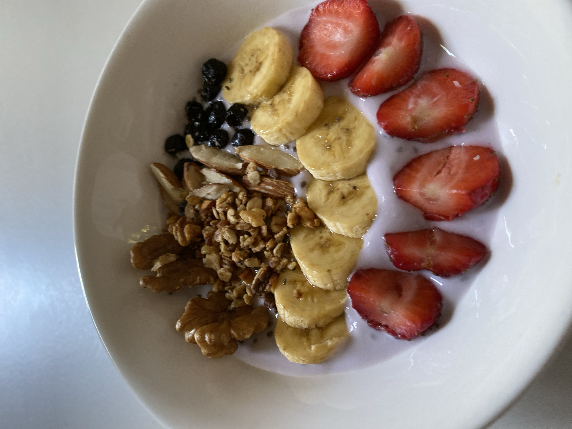 A white ceramic bowl contains a healthy breakfast dish. The dish consists of yogurt, topped with sliced bananas, halved strawberries, blueberries, granola, almonds, and walnuts. The meal is presented on a plain white surface, likely a table. The lighting suggests it is daytime. There is no indication of the specific location within Davao City, Philippines.