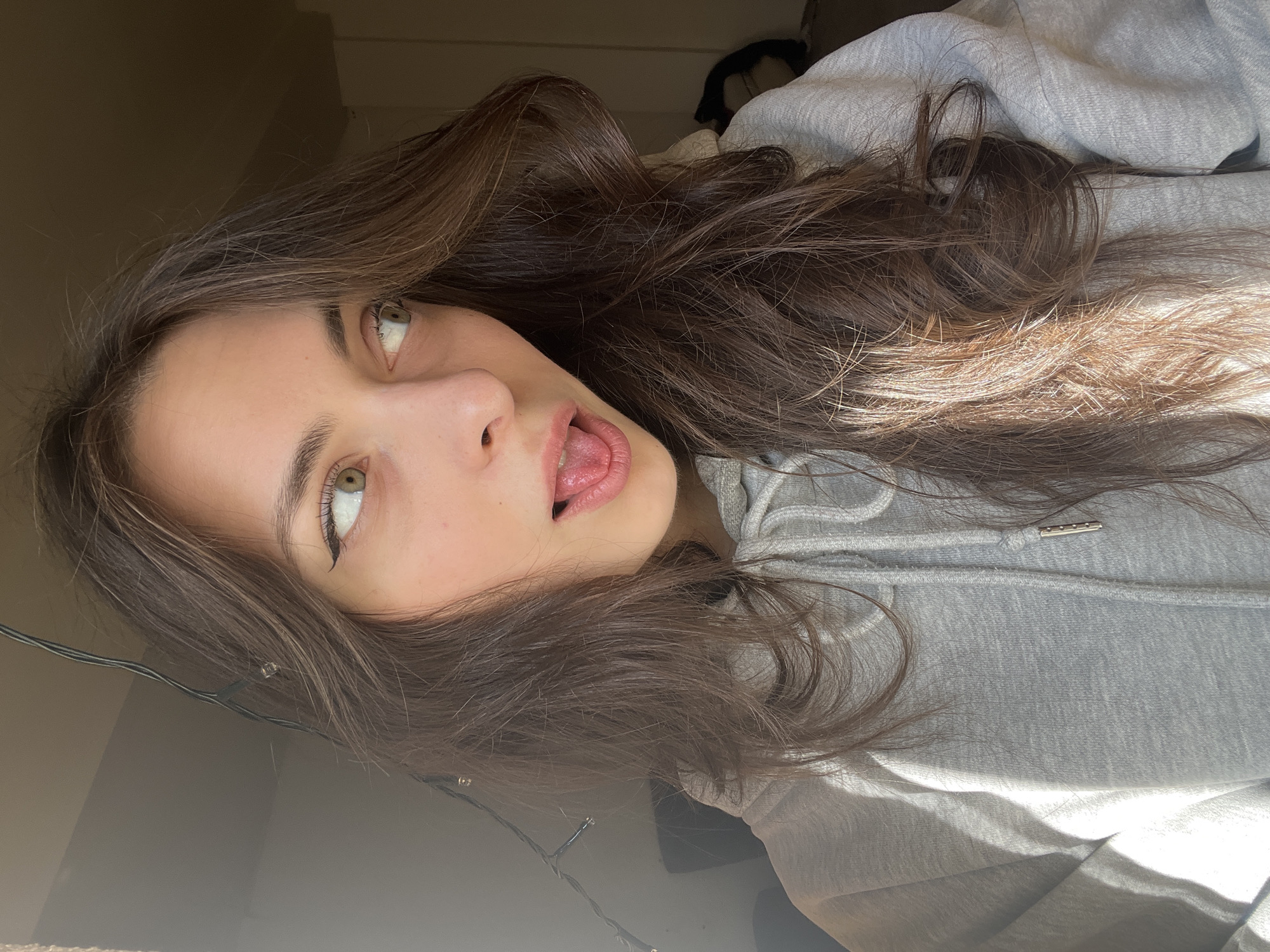 A young woman with long brown hair and hazel eyes is lying down, her head tilted to the side, with her mouth slightly open and her tongue visible. She is wearing a grey hooded sweatshirt. The background includes a wall, fairy lights and an indiscernible object. The location cannot be determined.